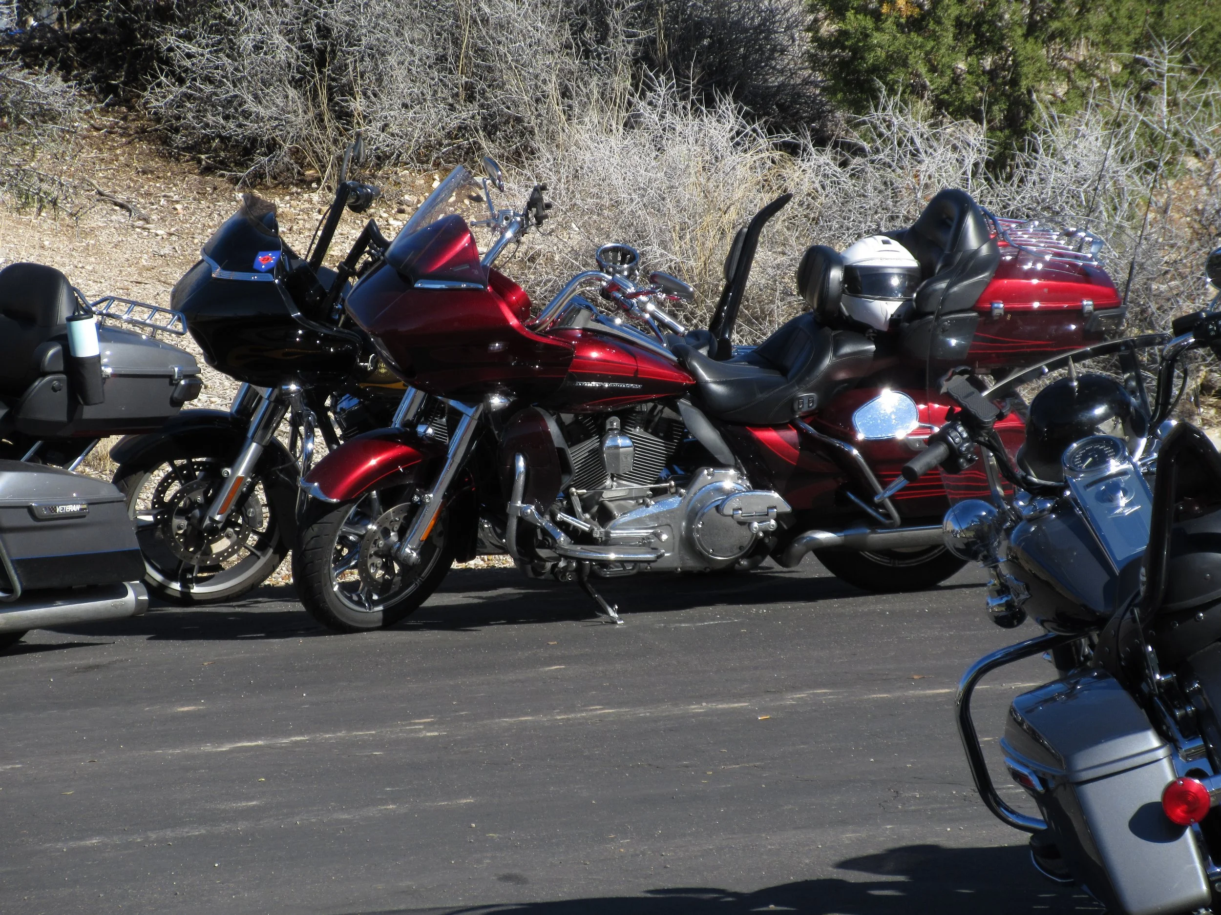Several motorcycles parked on a paved surface, with a red and black touring motorcycle in the center, surrounded by other motorcycles, in an outdoor area with dry bushes and trees in the background.