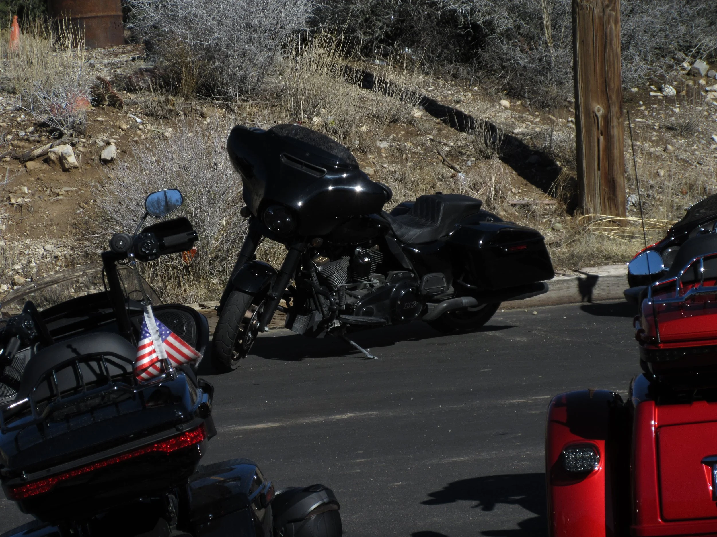 Black touring motorcycle parked on the side of a paved road, with dry bushes, rocks, and wood utility poles in the background.