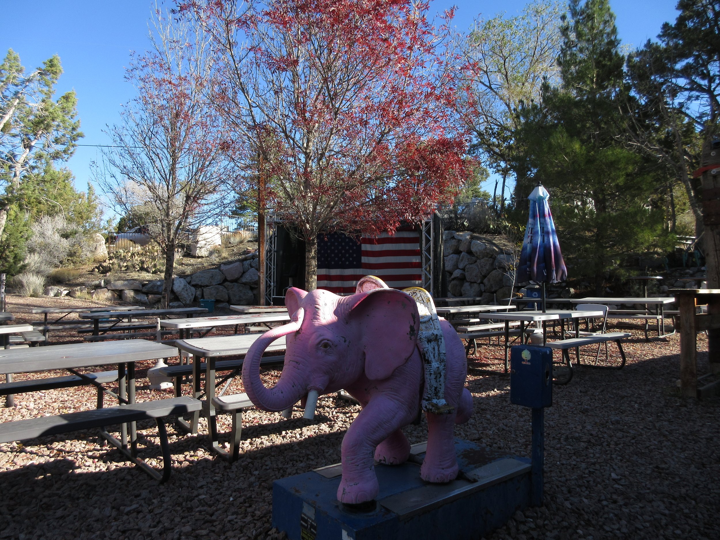 Pink elephant sculpture with a saddle in an outdoor setting, surrounded by picnic tables, trees, and an American flag backdrop under a clear blue sky.