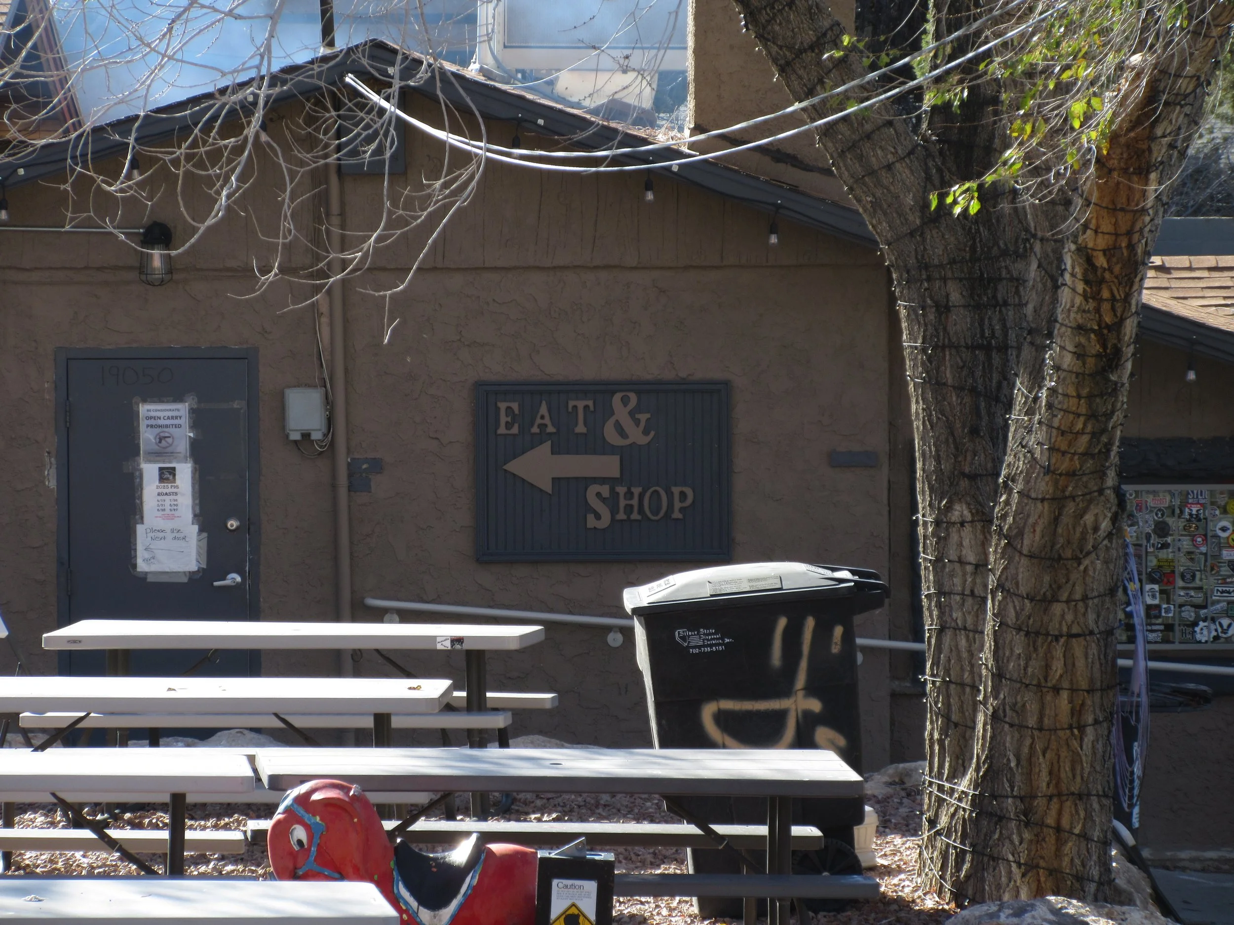 Building with a sign pointing to the left that reads 'EAT & SHOP,' surrounded by trees, tables, a trash bin, and picnic benches.