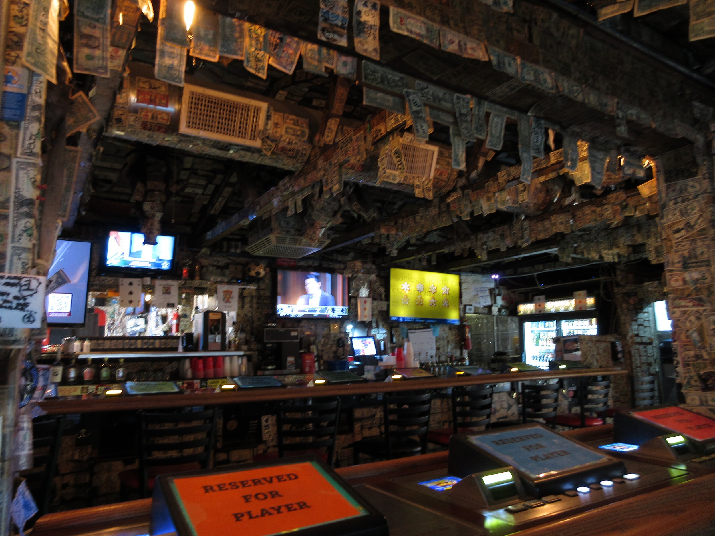 Interior of a bar with walls and ceiling covered in dollar bills, multiple TV screens, a long bar counter with reserved for player sign, and several chairs.