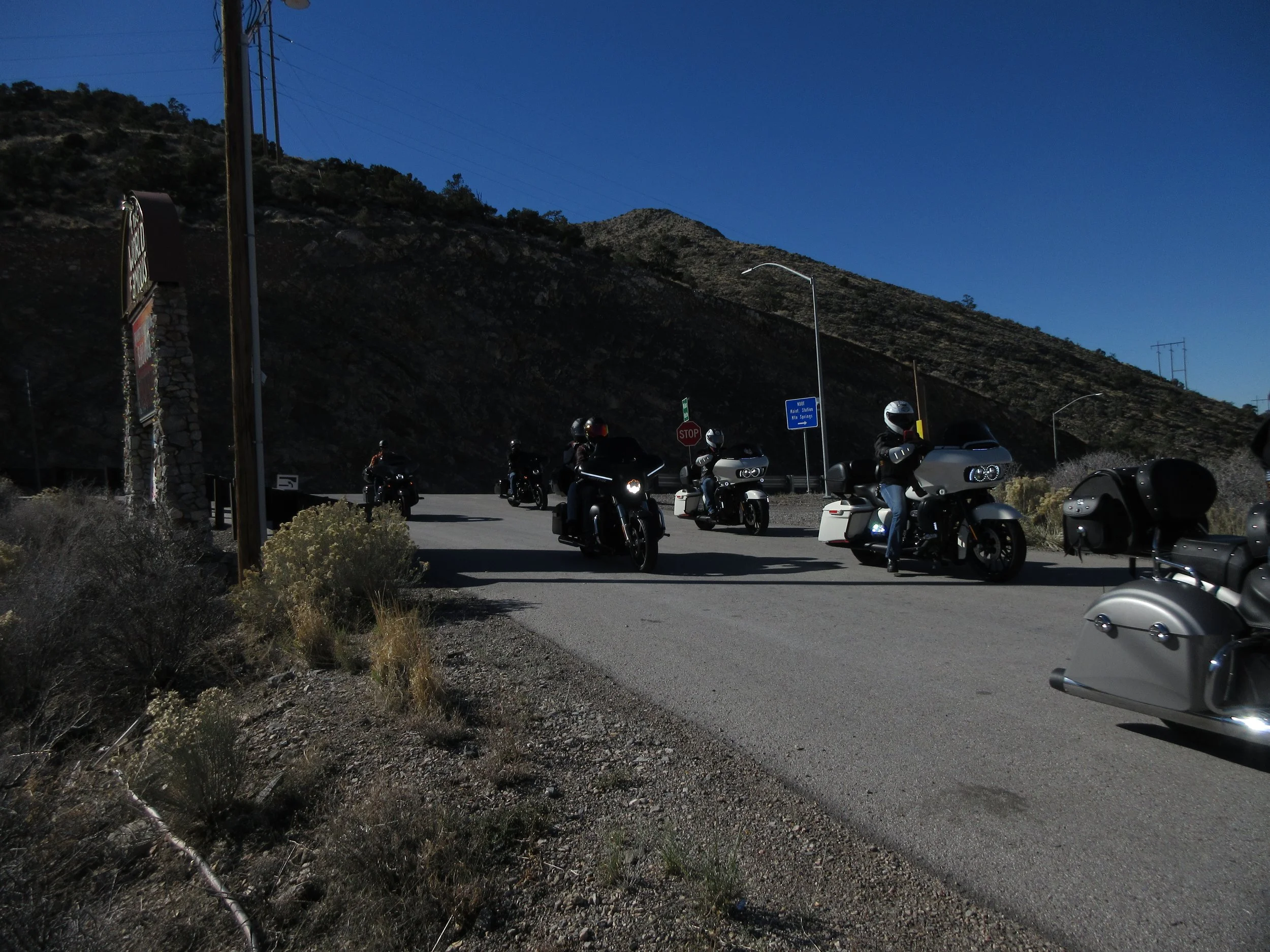 Group of motorcyclists riding on a mountain road with desert vegetation and mountainous terrain in the background.