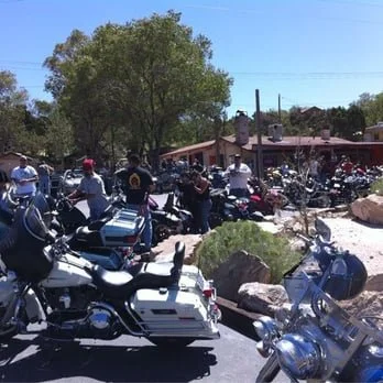 Multiple police motorcycles parked along a street with officers and bystanders nearby, trees and buildings in the background.
