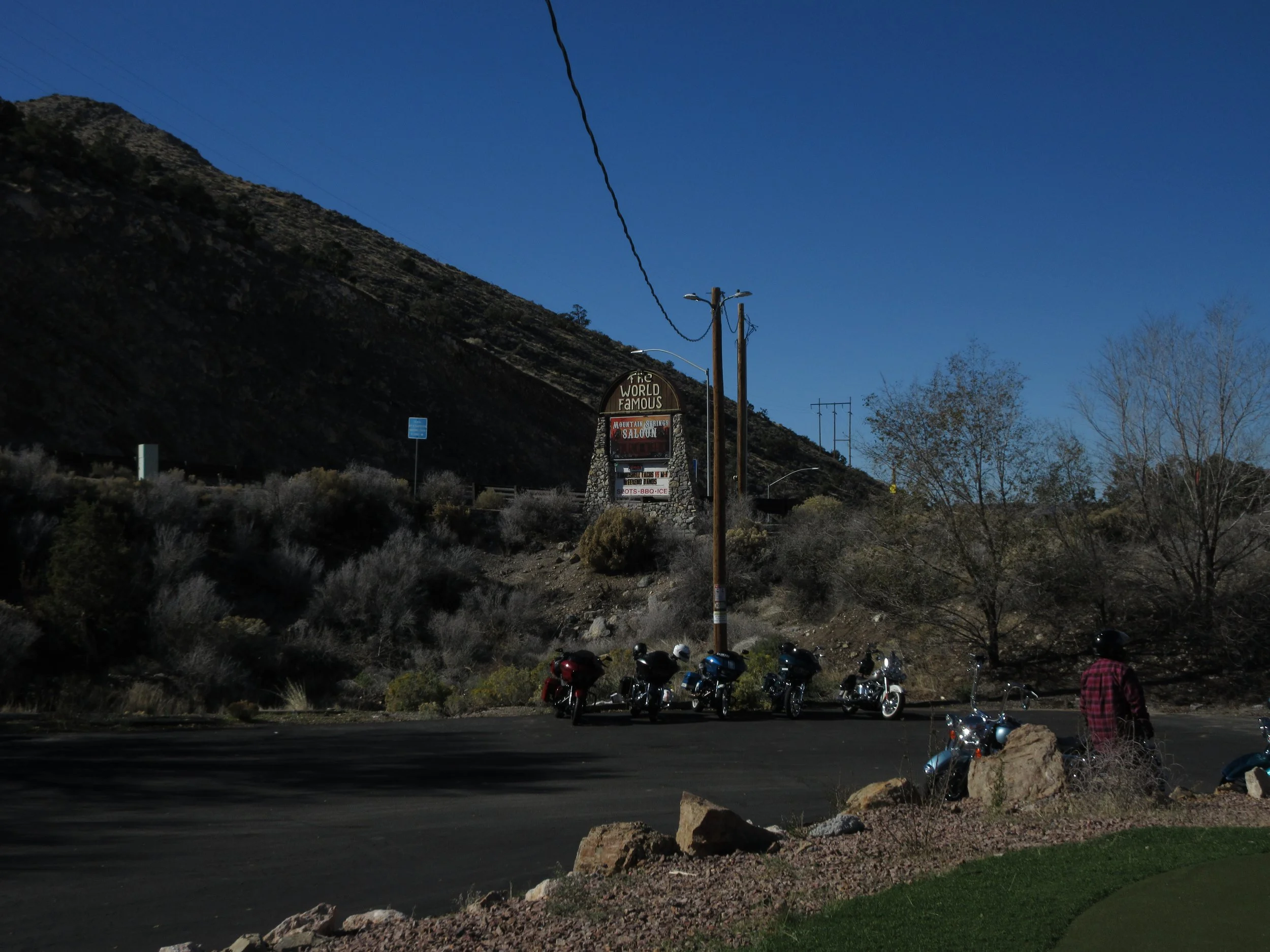 A parking lot with motorcycles parked along the curb, a person wearing a helmet and red checkered shirt standing next to a motorcycle, a large sign on a hillside in the background reading 'The World Famous Mountain Springs Saloon,' with a mountain an
