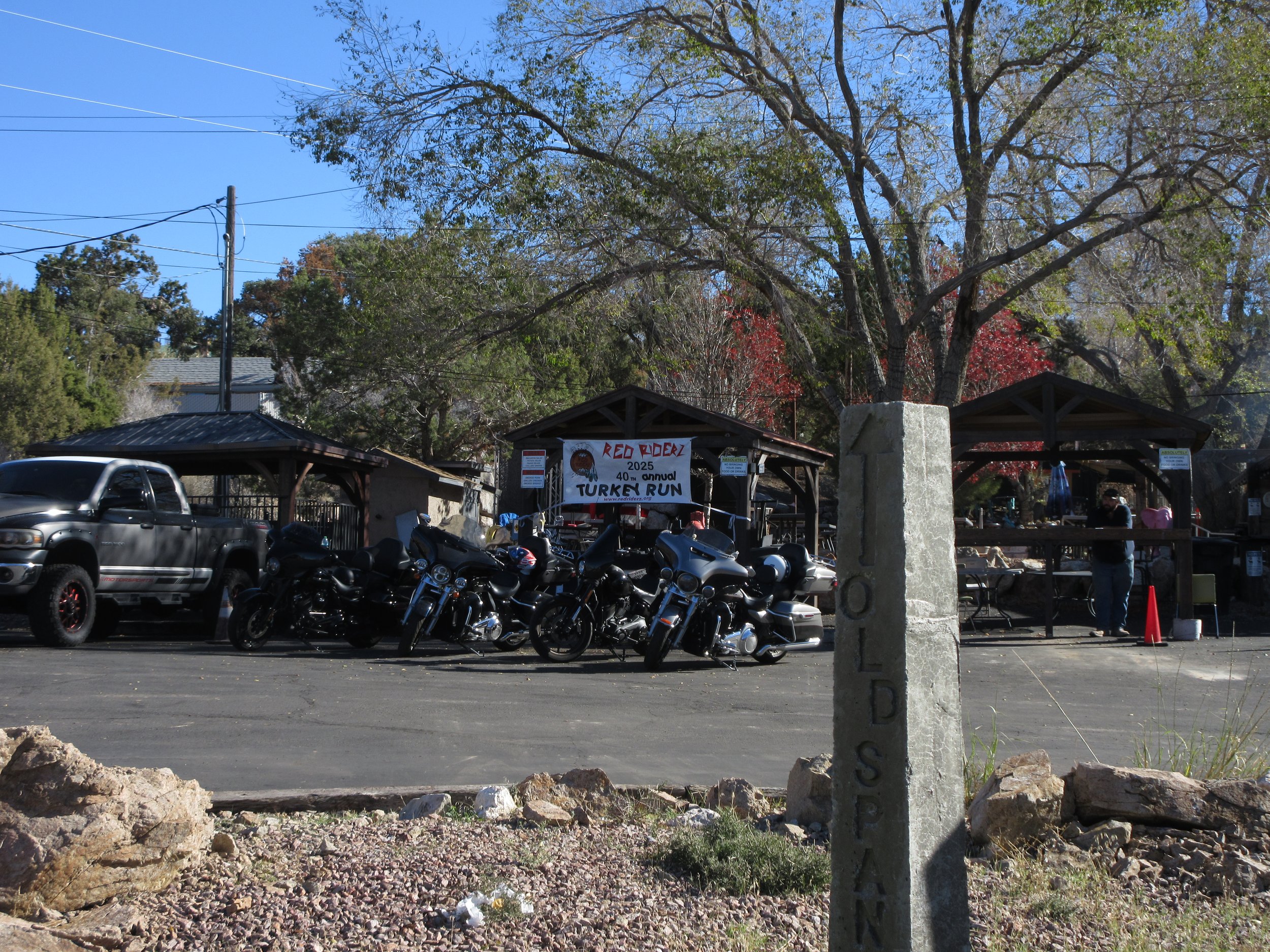 Outdoor scene showing a parking area with motorcycles and a pickup truck, a pavilion with a banner reading 'Red Riderz 2025 40th Annual Turkey Run,' trees with some red leaves, and a stone post with the word 'OLD SPAN' engraved. There are people sitt