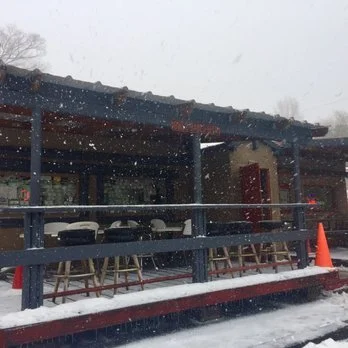 An outdoor patio of a cafe or restaurant during snowfall, with tables and chairs, a blue railing, and a traffic cone on the snow-covered ground.