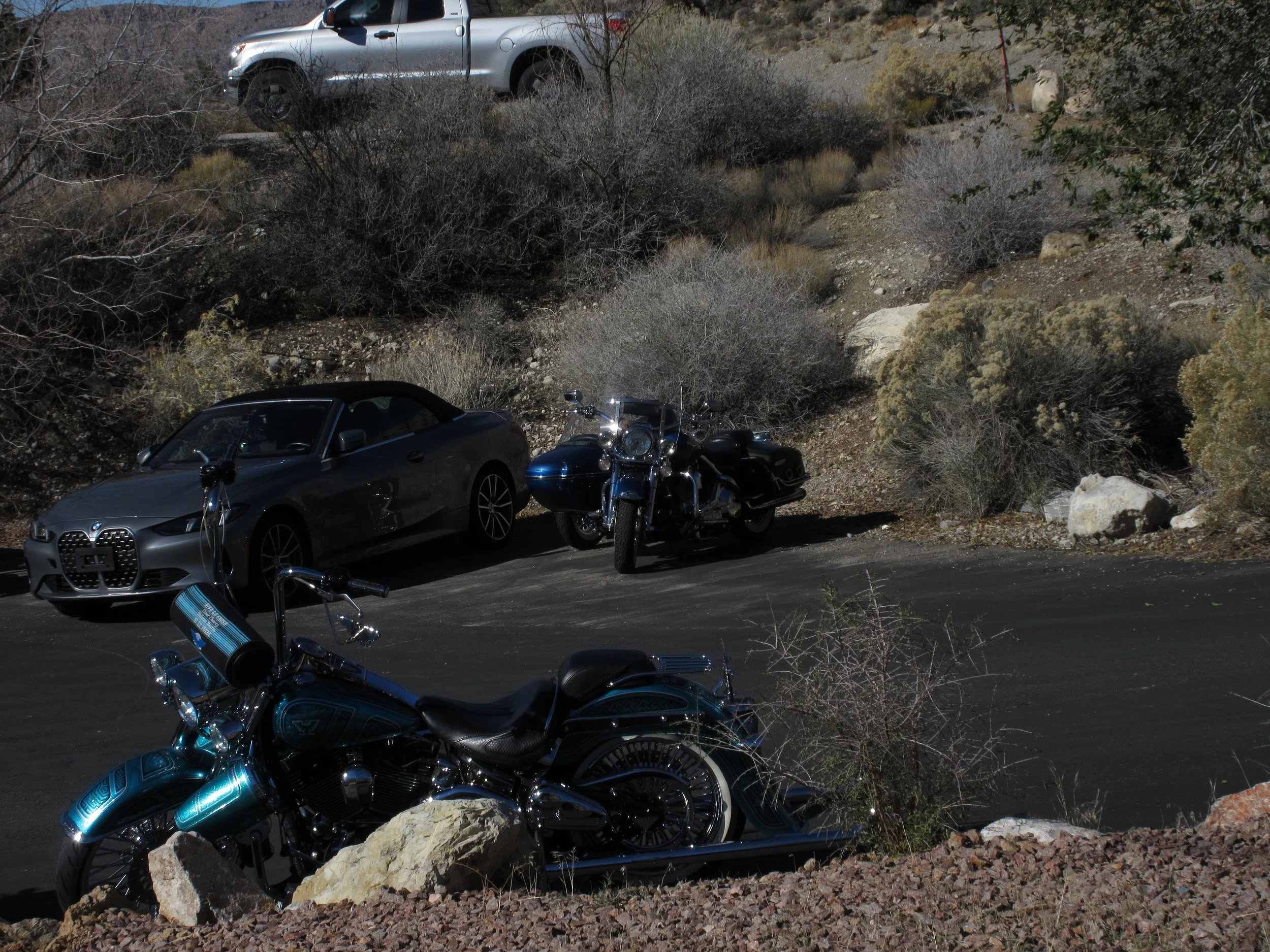 A parking area with two motorcycles, a car, and a motorcycle in the background, set in a desert-like landscape with dry bushes and rocks.