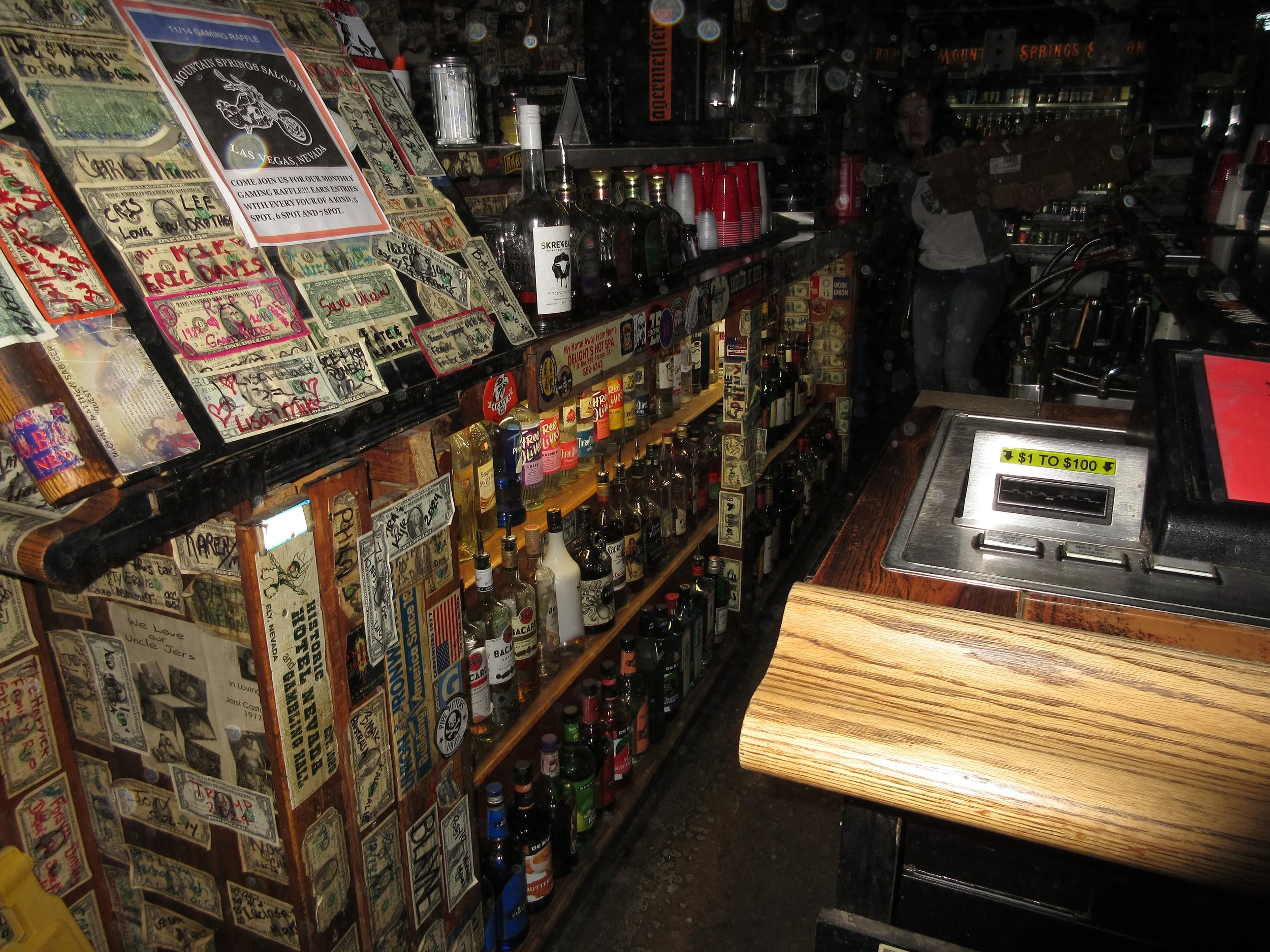 Inside a bar, shelves with various bottles of alcohol, signs, and stickers on the walls; a woman is standing behind the bar.