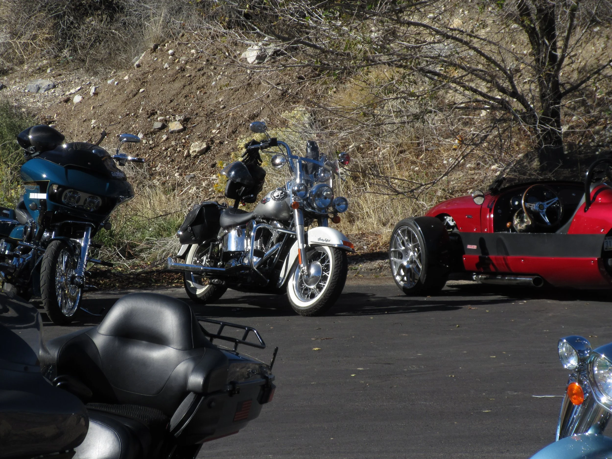 Several motorcycles and a red sports car parked on a paved area near a hillside with dry grass, rocks, and leafless trees.