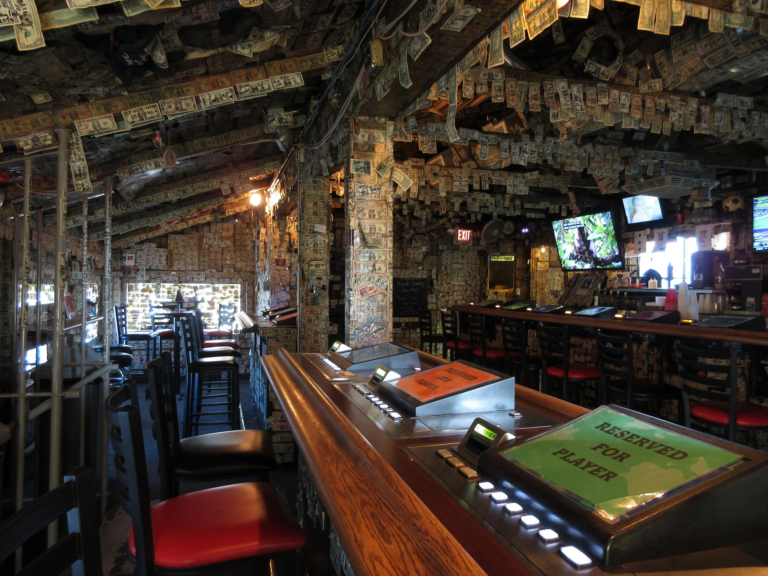 Inside a bar decorated with dollar bills covering the walls and ceiling, with bar stools, a counter with reserve signs, and multiple TV screens.