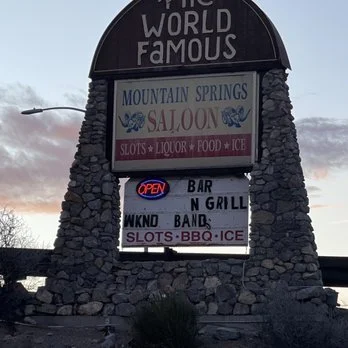 Signboard for a venue called Mountain Springs Saloon featuring slots, liquor, food, ice, and a bar and grill with karaoke and band, displaying an open sign.