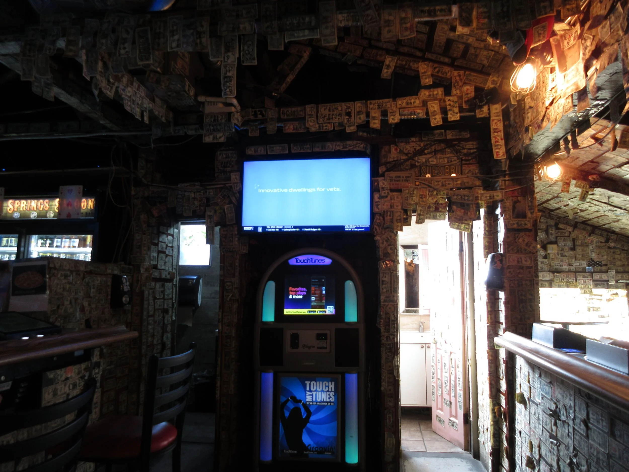 Interior of a bar or restaurant decorated with dollar bills covering the walls and ceiling. There is a TouchTunes jukebox in the center and a large flat screen TV on the wall displaying a blue screen with text.