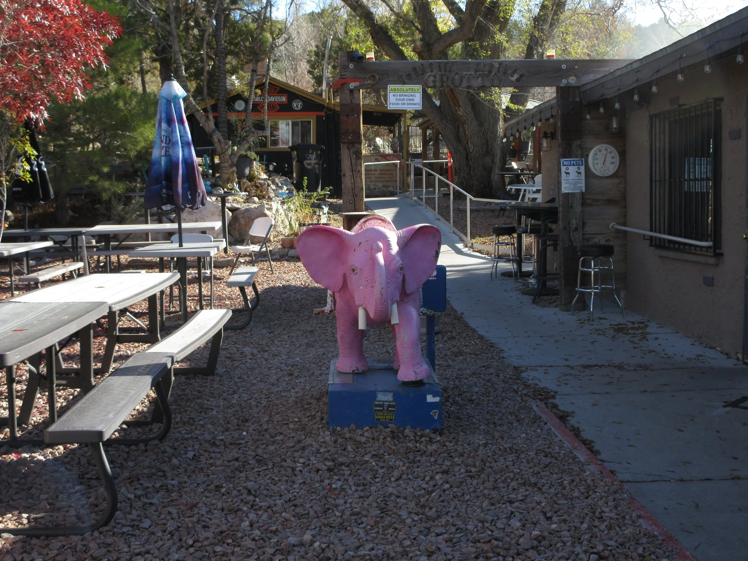 Pink elephant sculpture on a blue base outside a rustic restaurant with outdoor seating and gravel path.