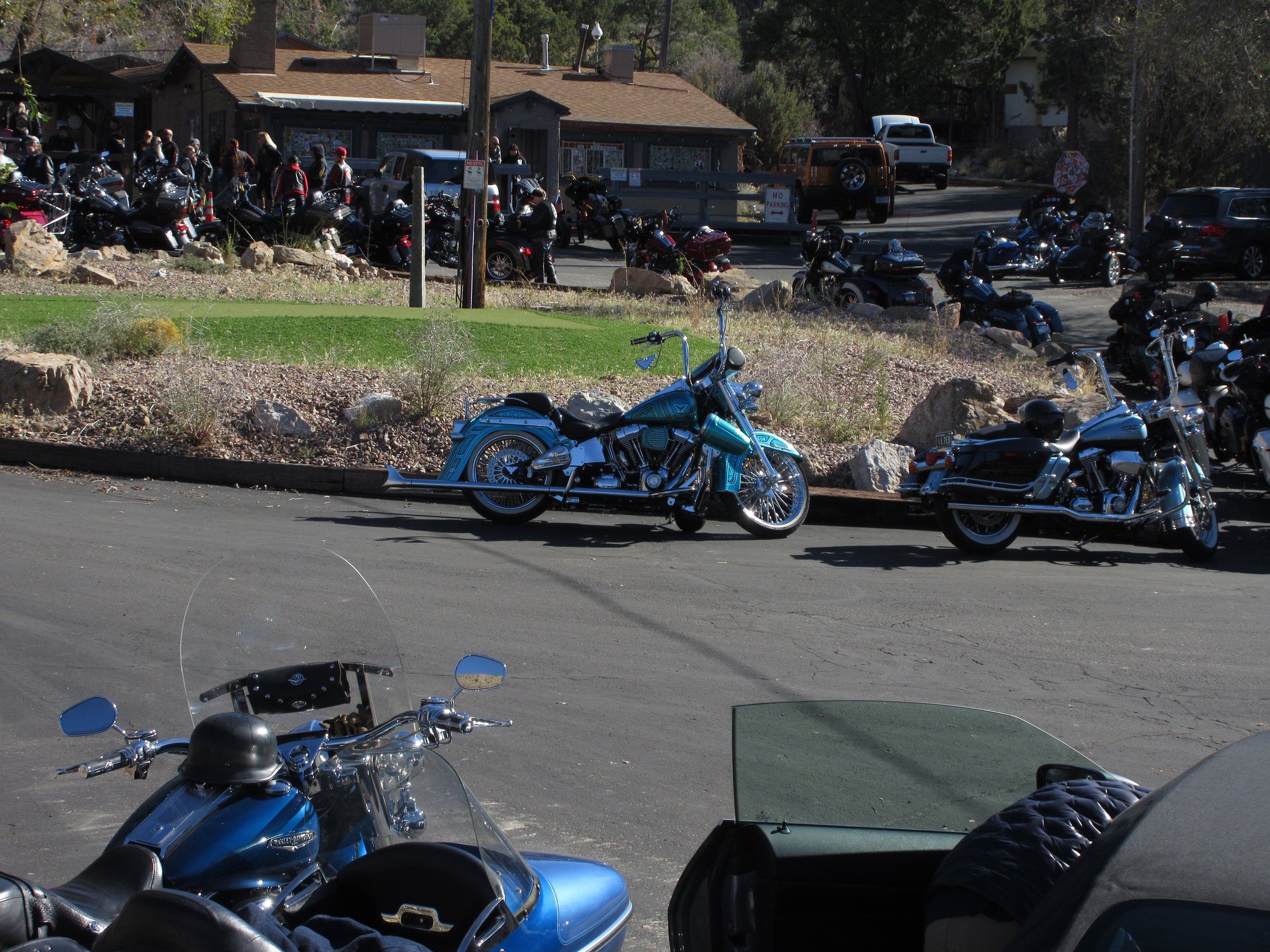 A parking lot with multiple parked motorcycles and a few cars, with a building and people in the background.
