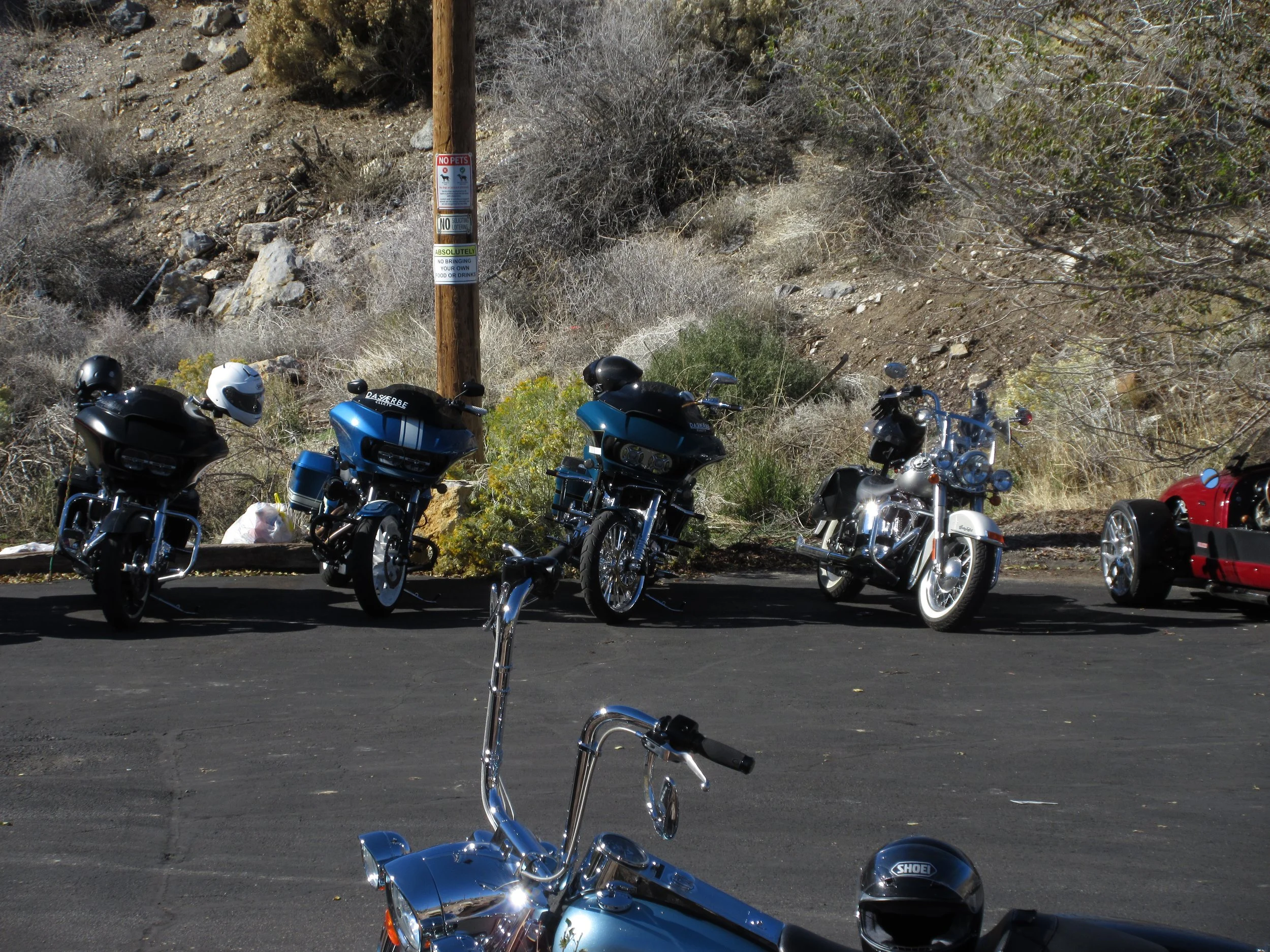 Five motorcycles parked on black asphalt in front of a rocky hillside with bushes, a wooden pole with signs, and a clear sky.