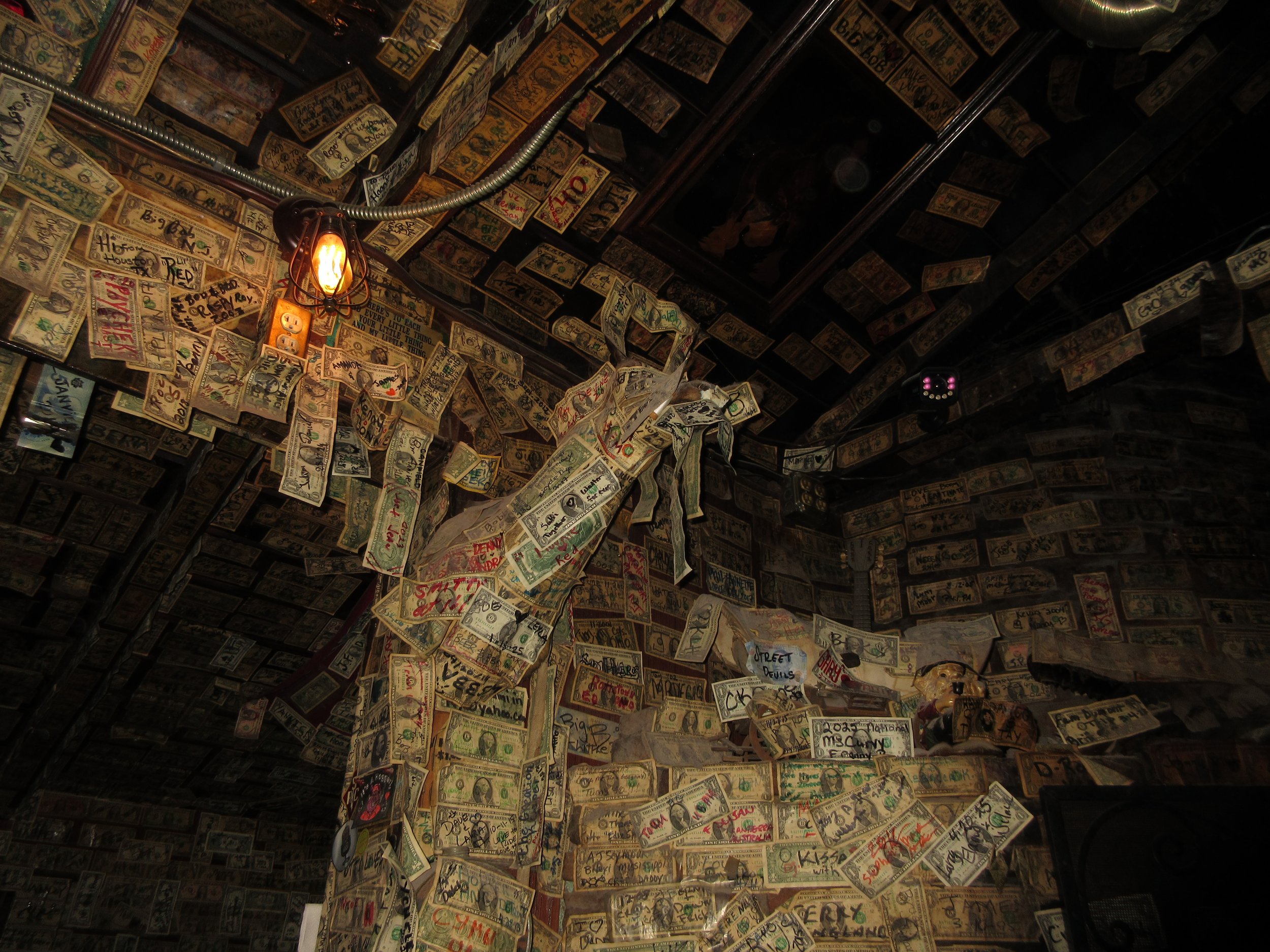 Interior of a room decorated with numerous hanging and taped American dollar bills on the walls and ceiling, with a visible light bulb hanging from the ceiling.