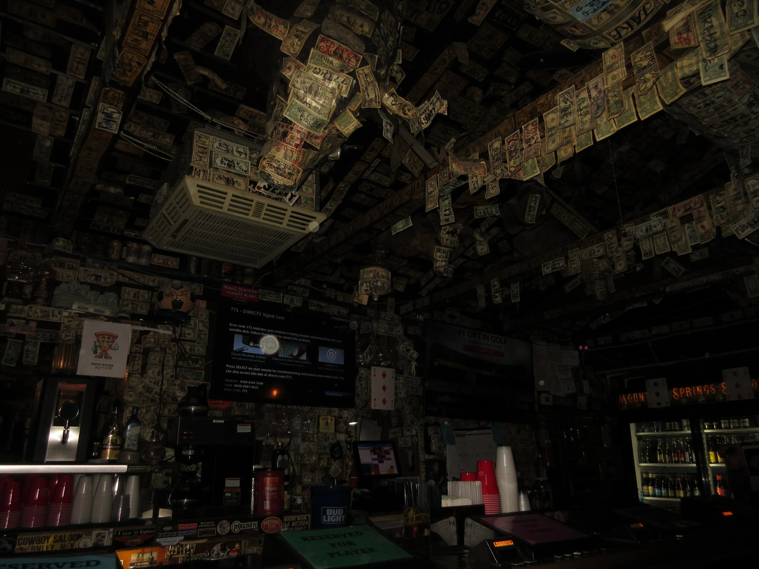 Bar interior heavily decorated with hanging dollar bills covering ceiling and walls, with various signs, a television, and beverage refrigerators.