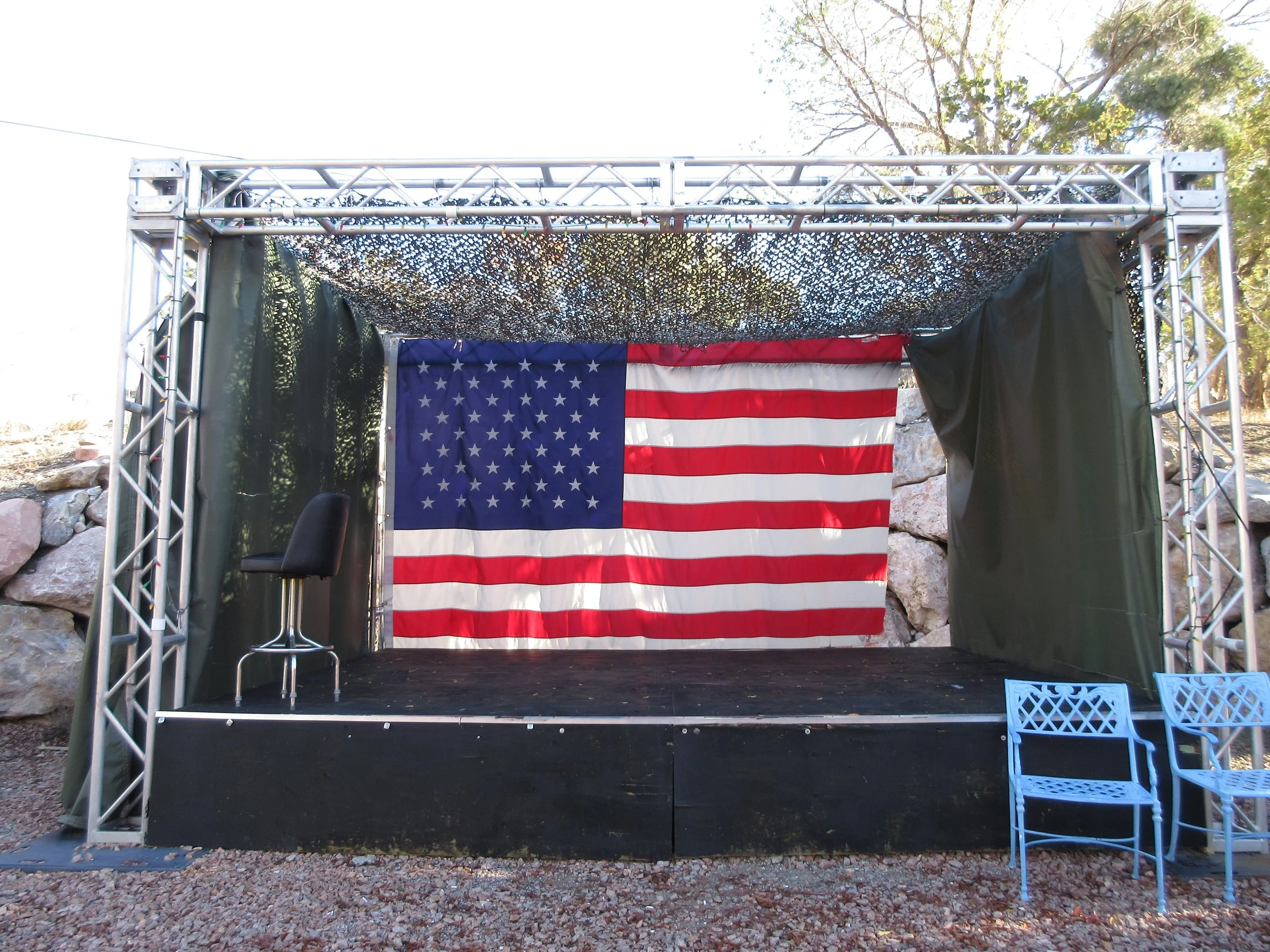 An outdoor stage with an American flag hanging at the back, a black chair on the left side, and two blue chairs on the right side, with trees and rocks in the background.