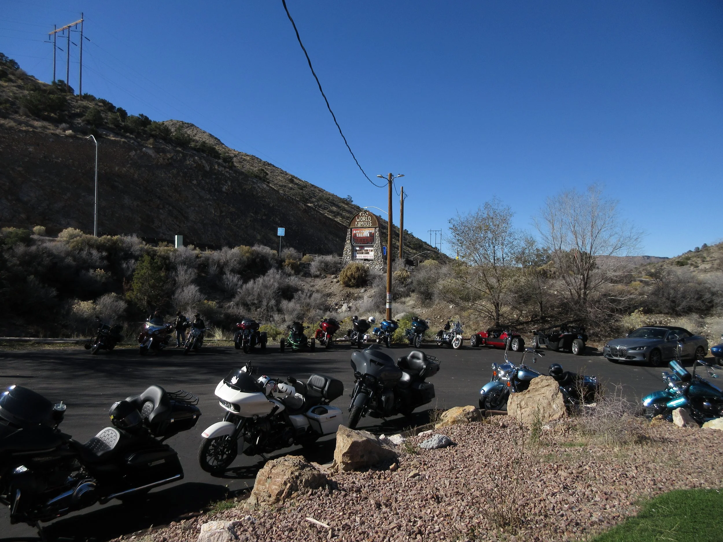Parking lot with motorcycles and cars against a desert hillside, with a sign and power lines, under a clear blue sky.