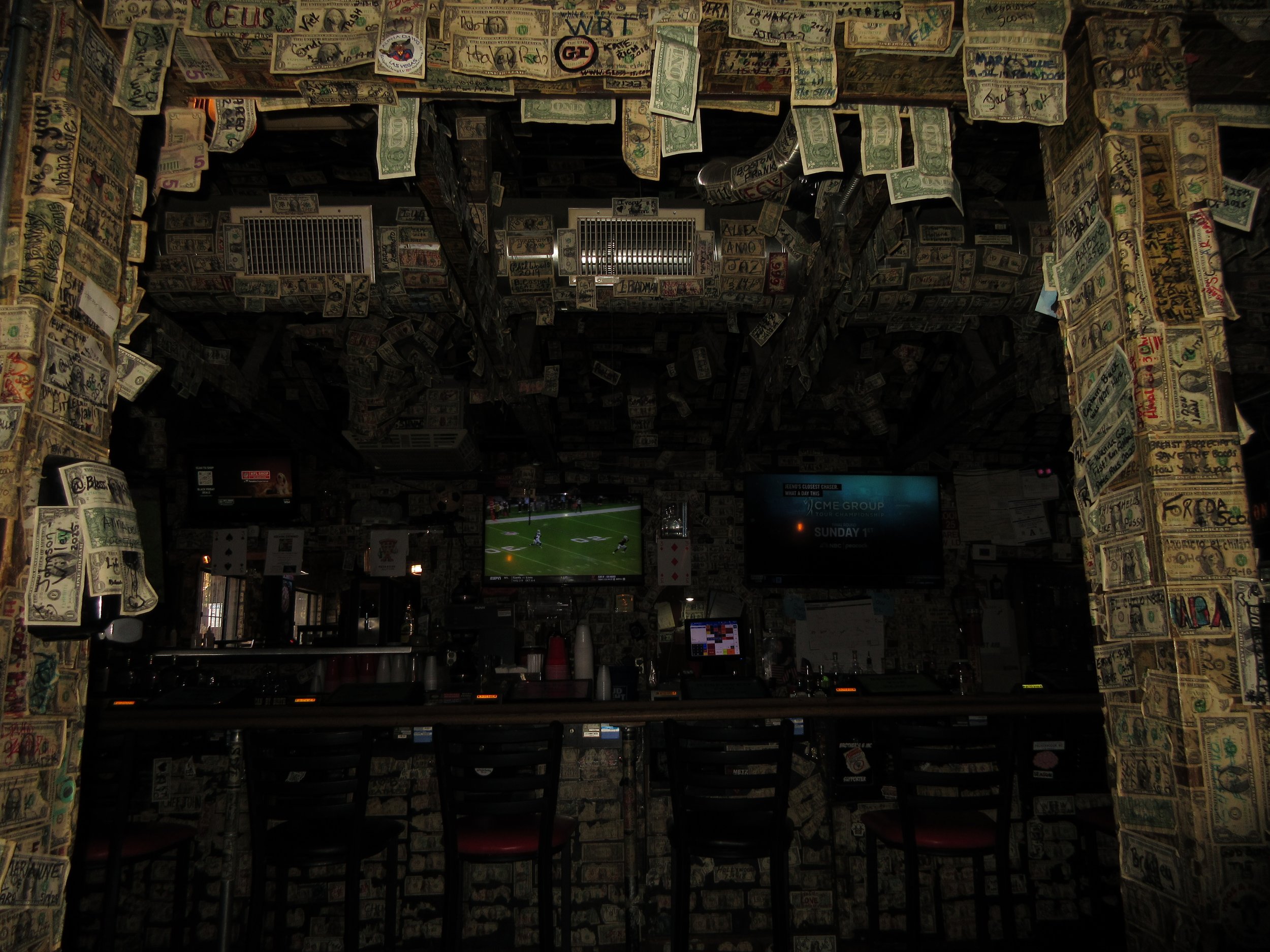 Bar interior covered in dollar bills with televisions on the wall showing sports games.