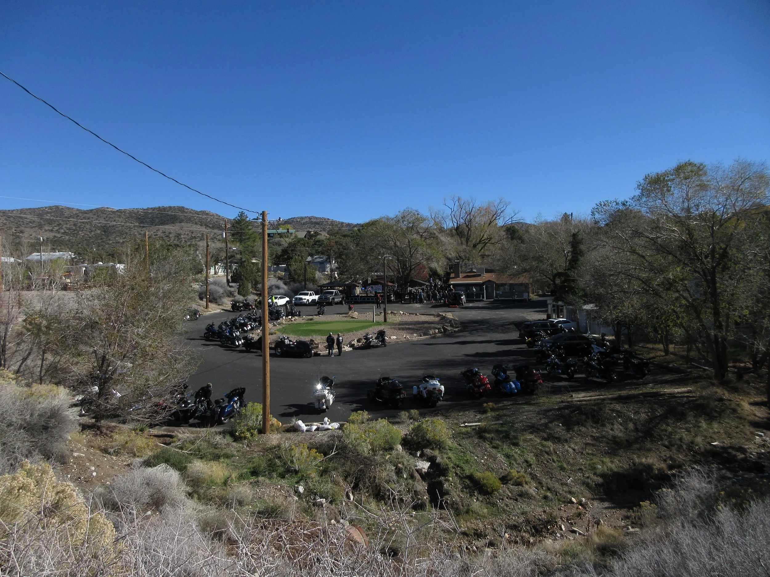 A parking lot with motorcycles and cars, a group of people gathered, and buildings with trees in the background under a clear blue sky.