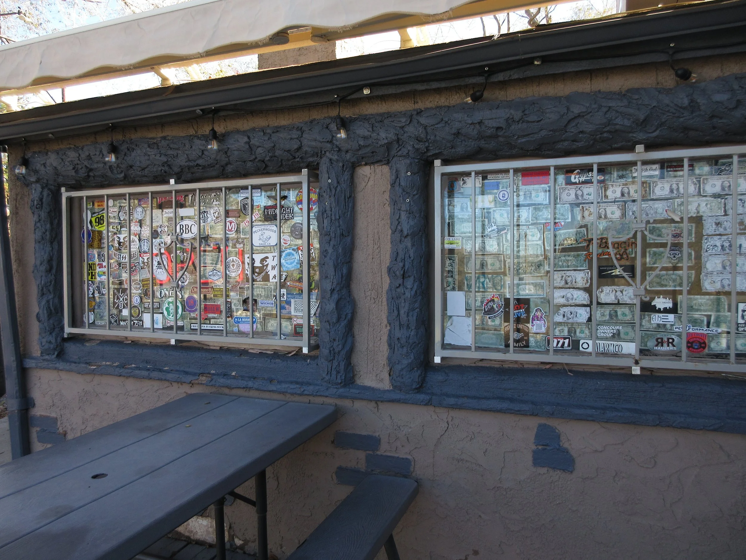 Two windows with metal bars on a building, covered with various stickers and decals. An outdoor wooden bench is in front of the windows.