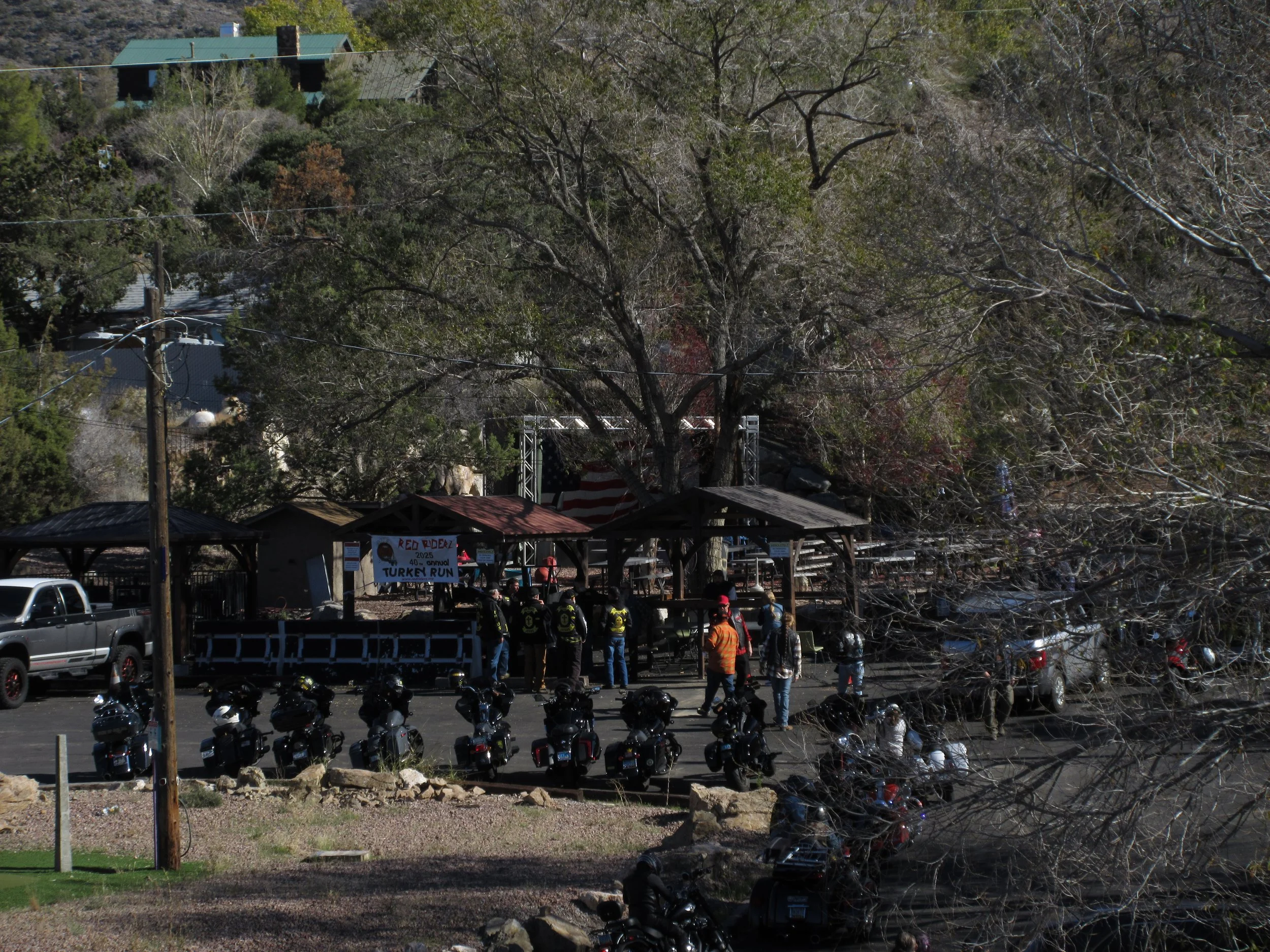 A gathering of people and motorcycles in a parking lot near a small stage with banners for a turkey run event, surrounded by trees and outdoor structures.