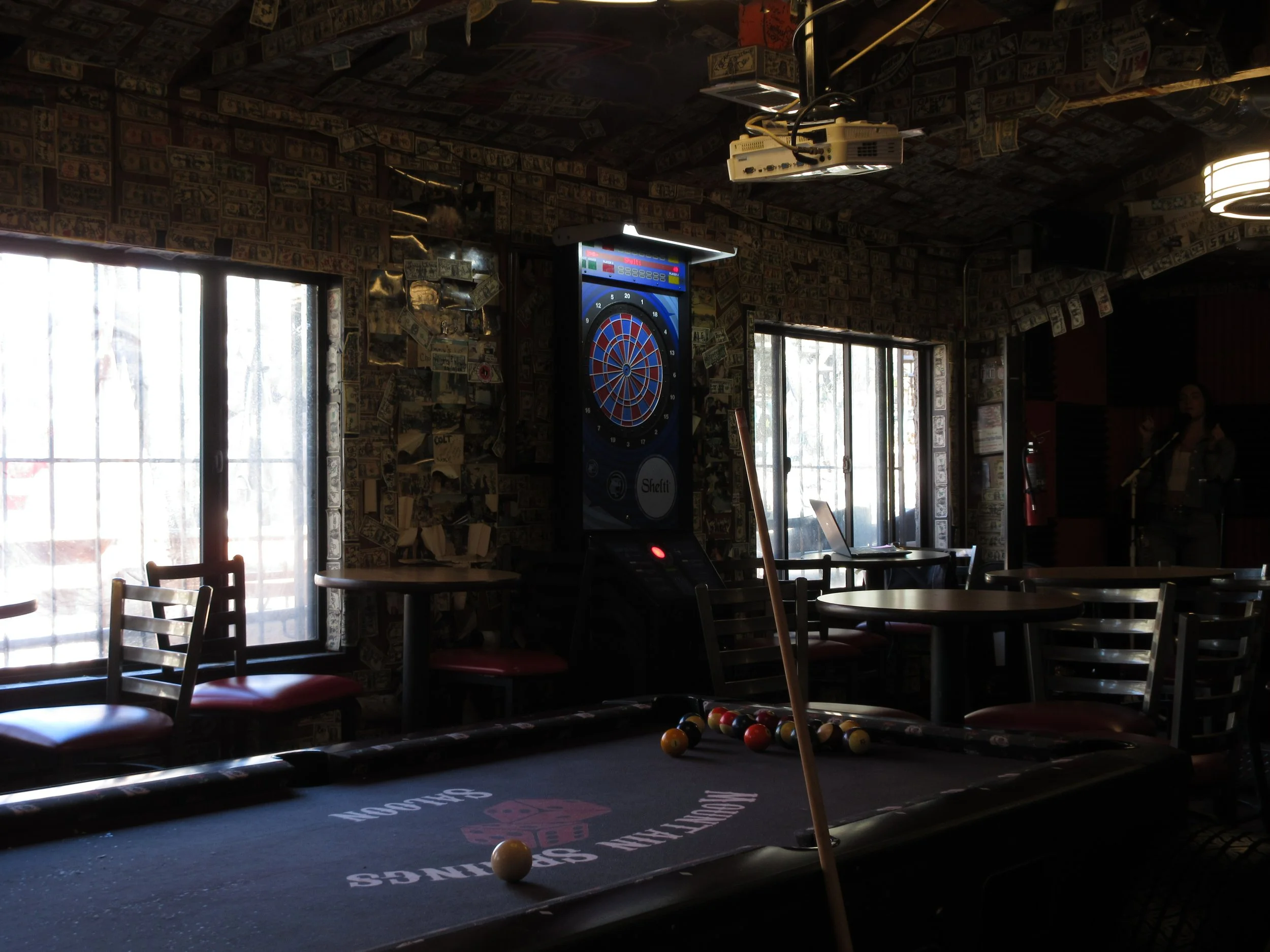Dimly lit bar or recreational room with a pool table in the foreground and a dartboard on the wall. Windows let in natural light, creating a cozy atmosphere. There are chairs and tables around, and a person is standing near a microphone on the right 