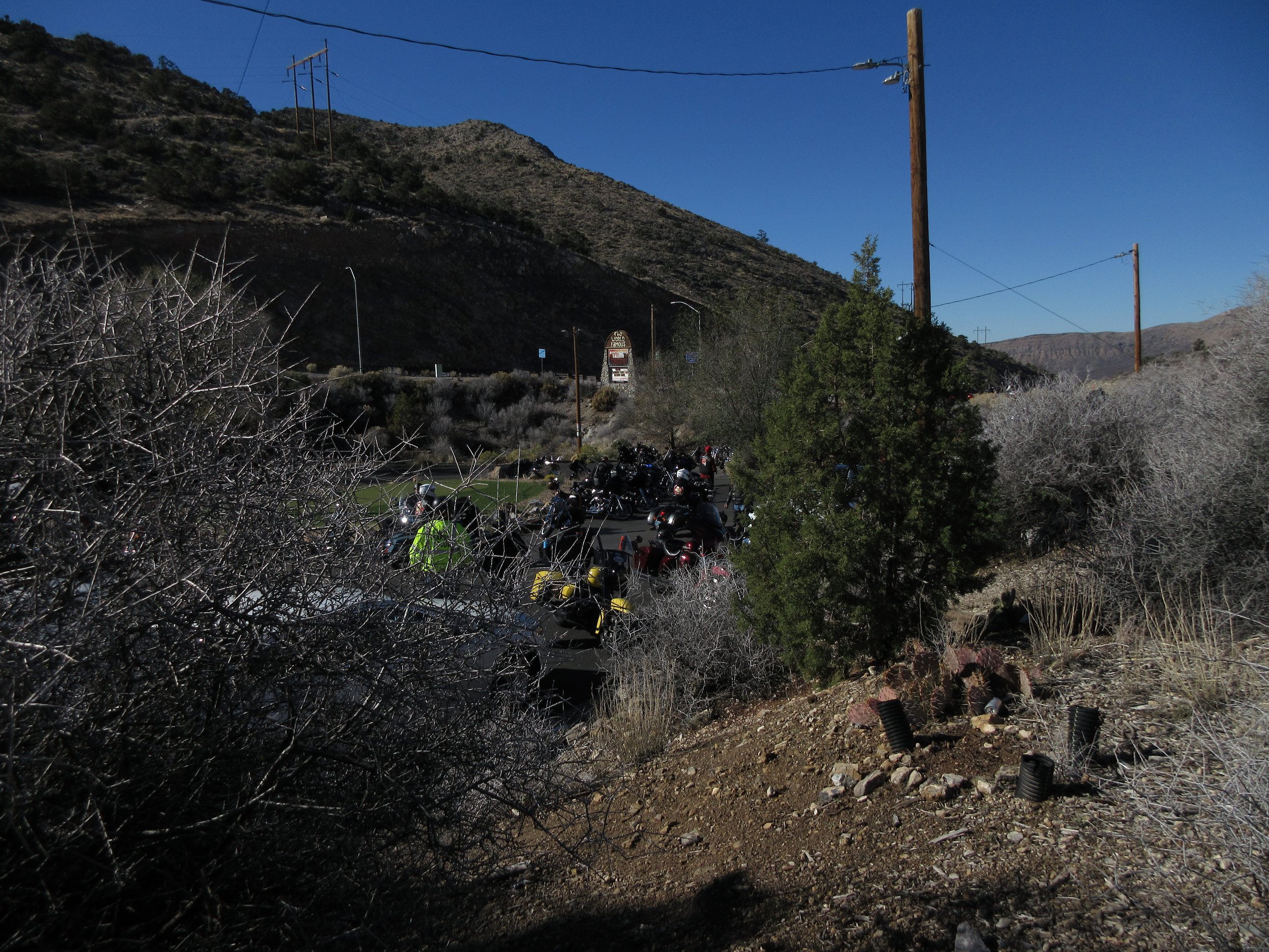 Parking lot filled with motorcycles, surrounded by dry bushes and trees, with hills and utility poles in the background under a clear blue sky.