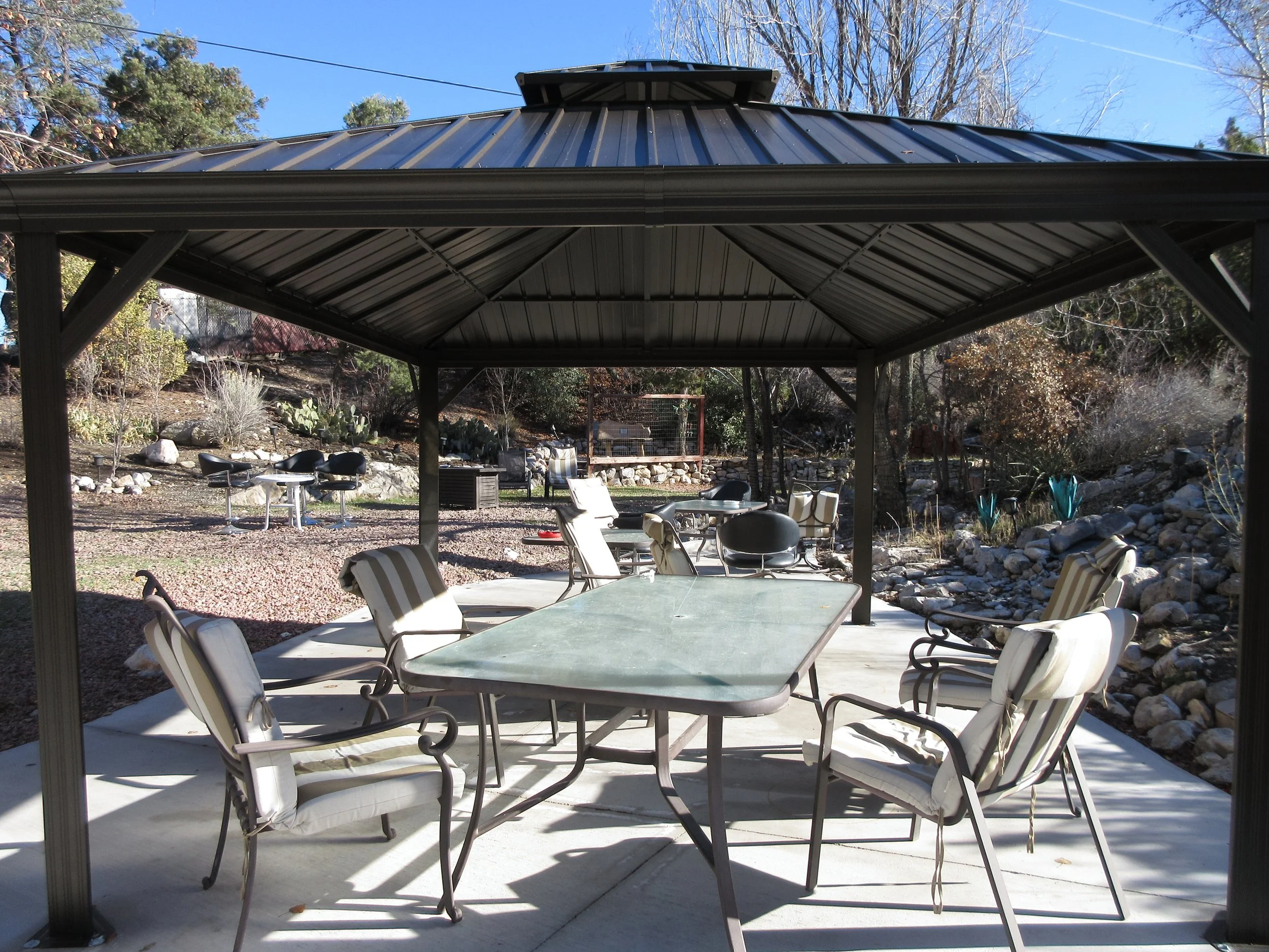 Outdoor patio with a metal roof shelter, a large rectangular table, and surrounded by chairs, with a landscaped yard featuring rocks, plants, and trees in the background.