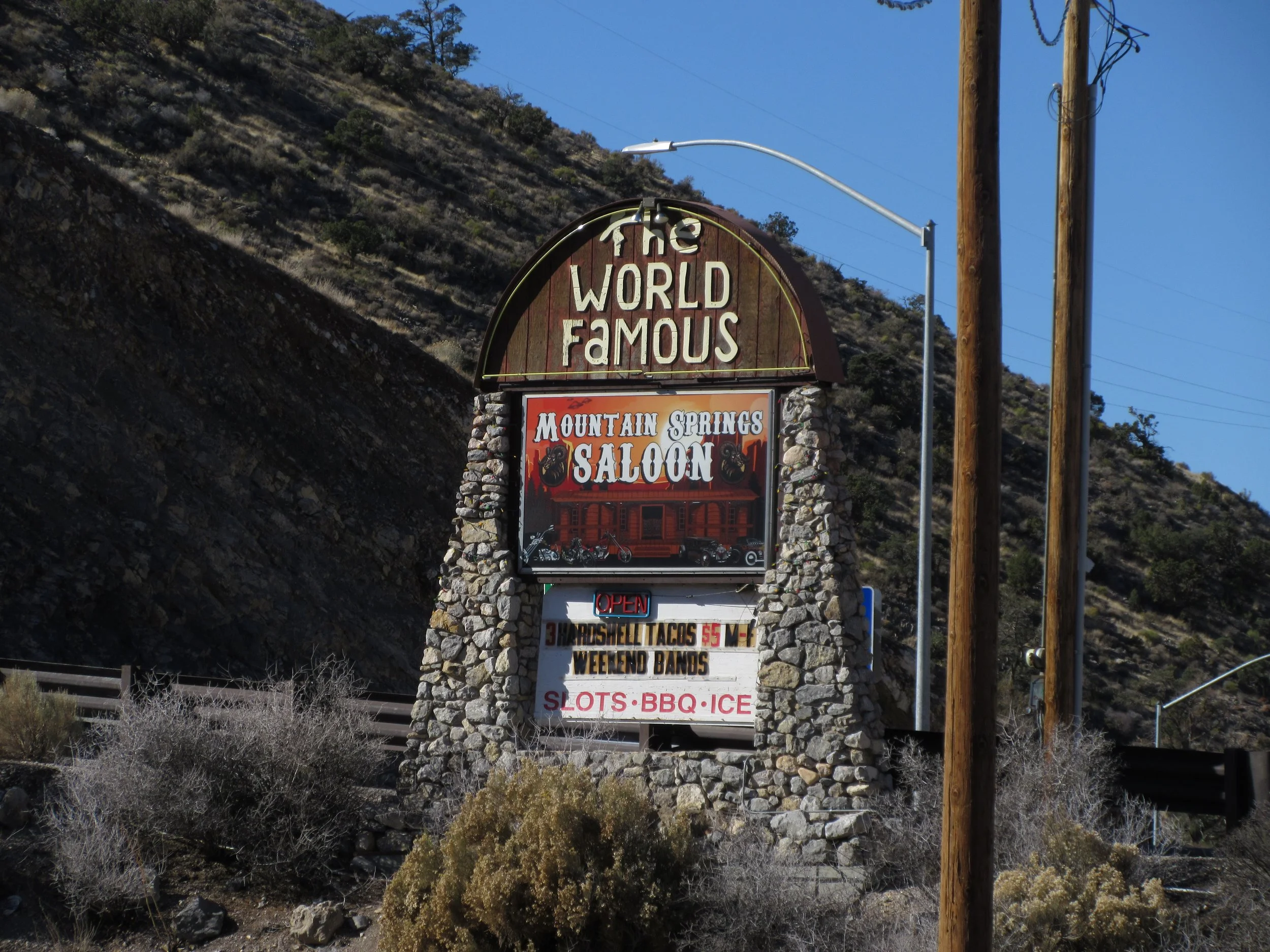 Sign for The World Famous Mountain Springs Saloon with details of food and entertainment options on a stone structure, surrounded by desert bushes and hills under a clear blue sky.