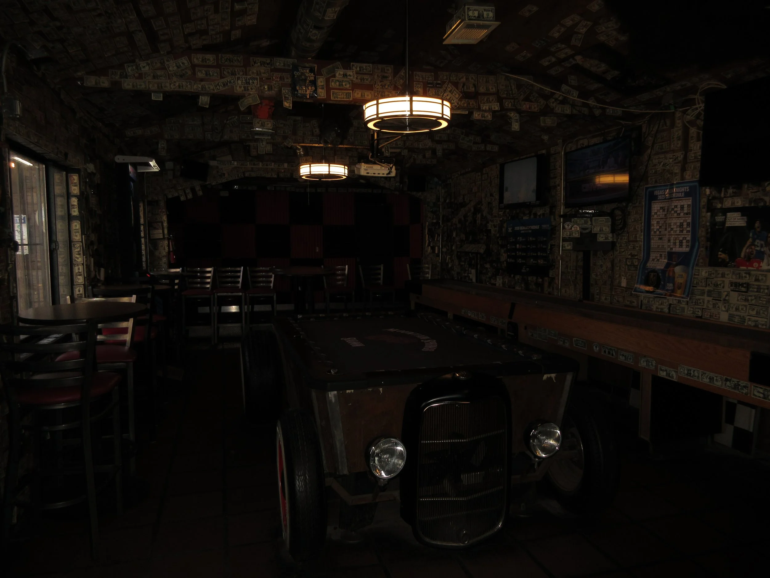 Dimly lit bar or entertainment venue with a car-shaped game table in the foreground, walls and ceiling decorated with dollar bills, flat-screen TVs, posters, and framed pictures, with some tables and chairs visible.