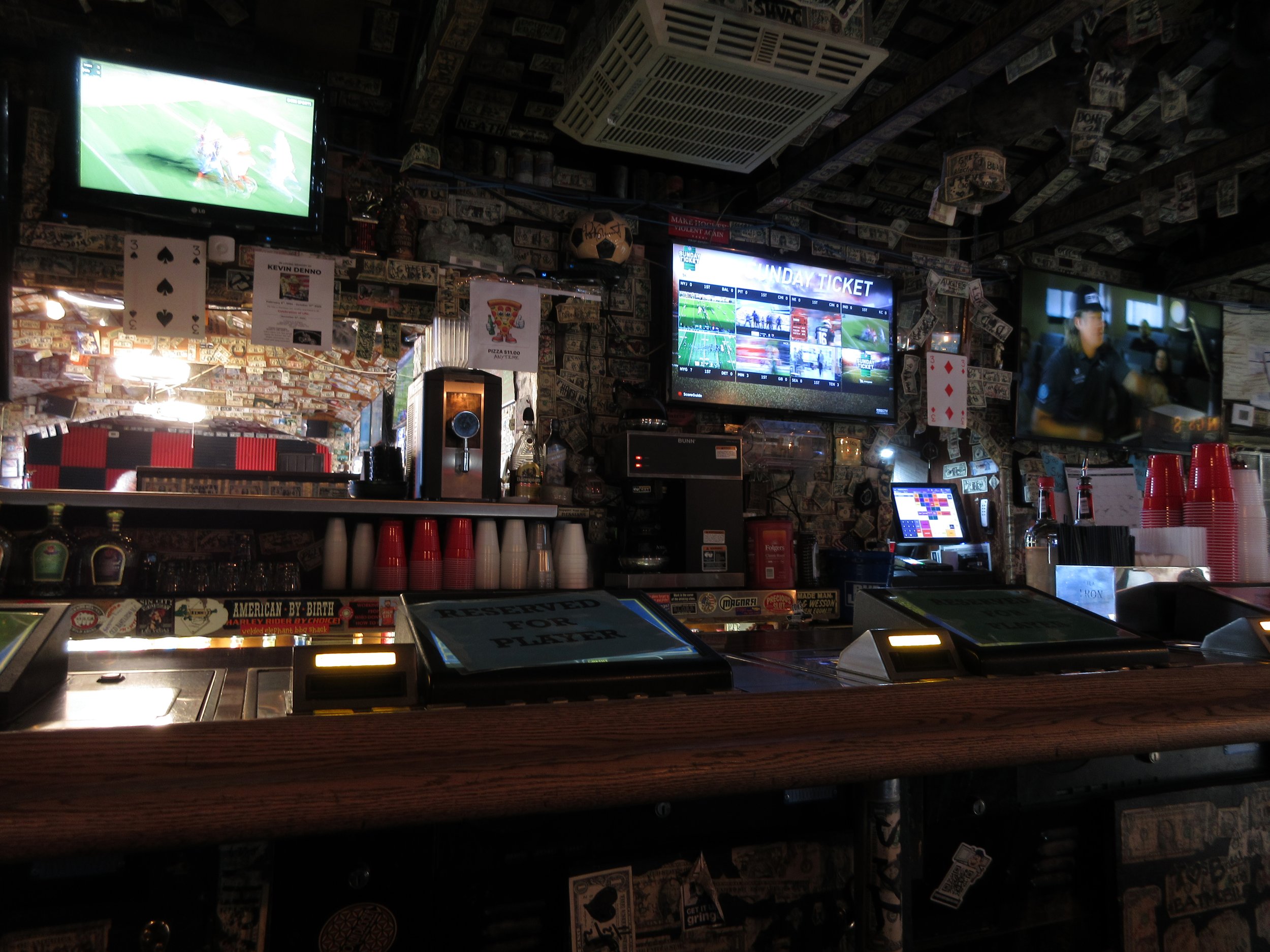 A bar with multiple television screens showing sports and a card game on the wall. The bar counter has red cups, a coffee machine, and menu signs. The walls are decorated with dollar bills and playing cards.
