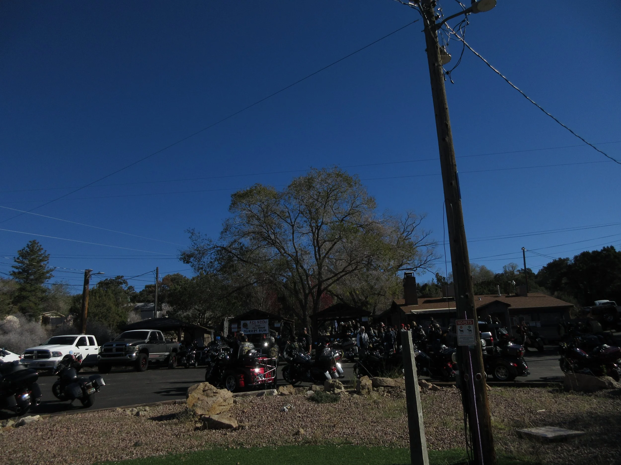 Parking lot filled with motorcycles and trucks with a building in the background, trees, power lines, and a clear blue sky.