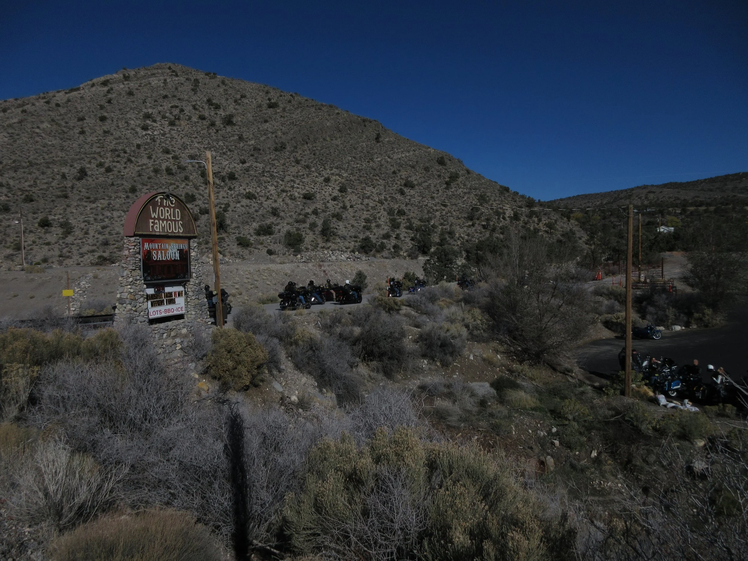 Sign for The World Famous Mountain Springs Saloon with a mountainous desert landscape and parked motorcycles in the background.