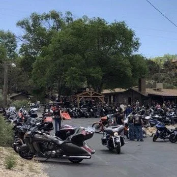A busy parking lot filled with motorcycles and a few cars, with people walking around near a wooden structure and trees in the background.