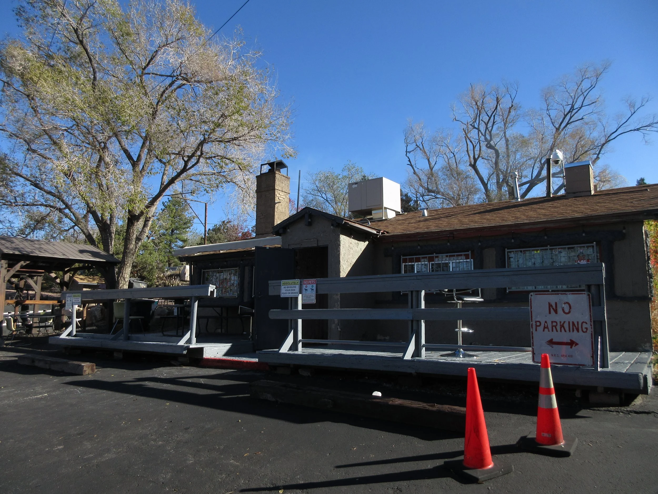 A small building with a deck and caution cones in front, surrounded by leafless trees against a clear blue sky.
