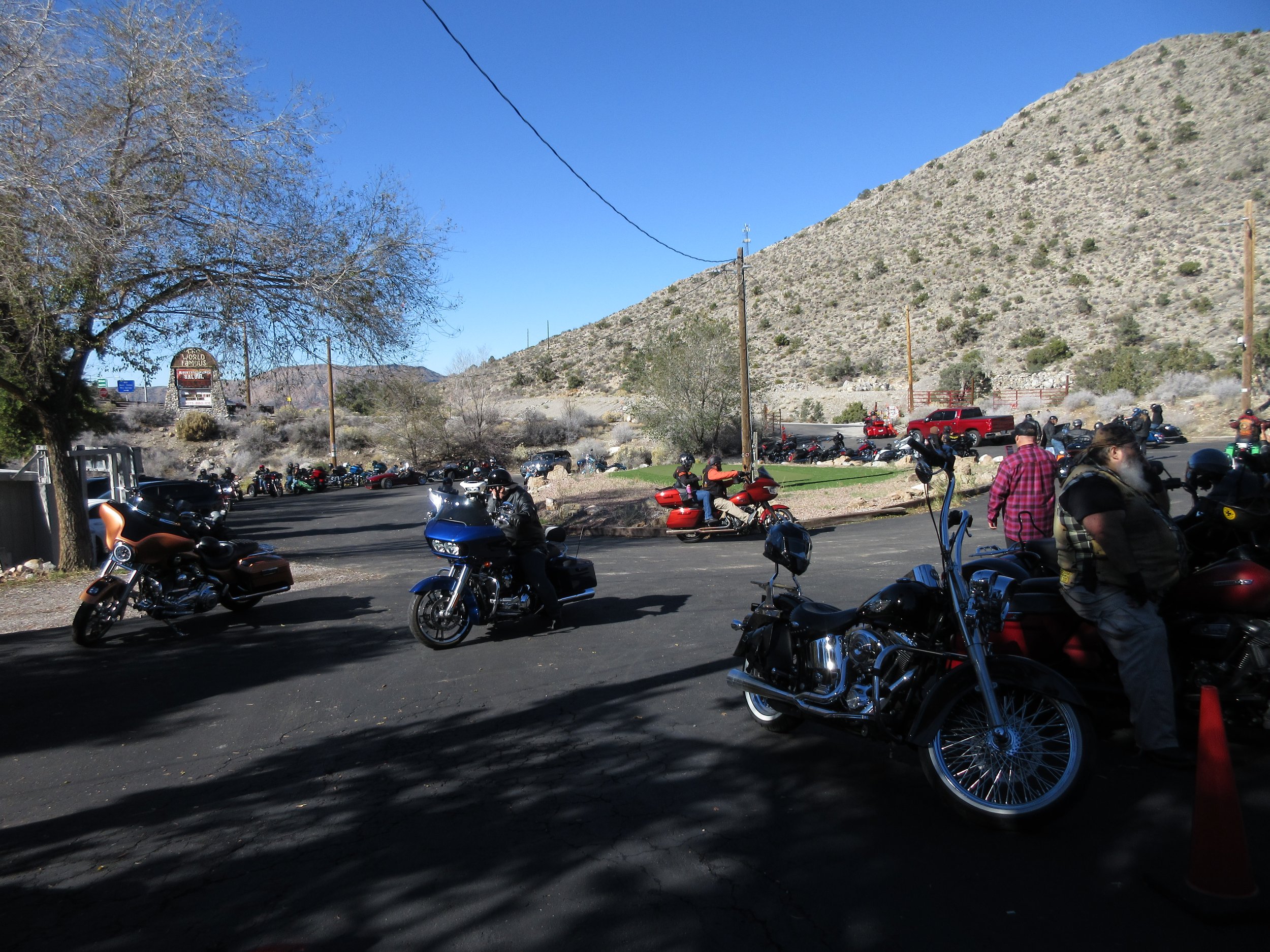Motorcycles parked in an outdoor lot with people standing nearby, in a mountainous area under clear blue skies.