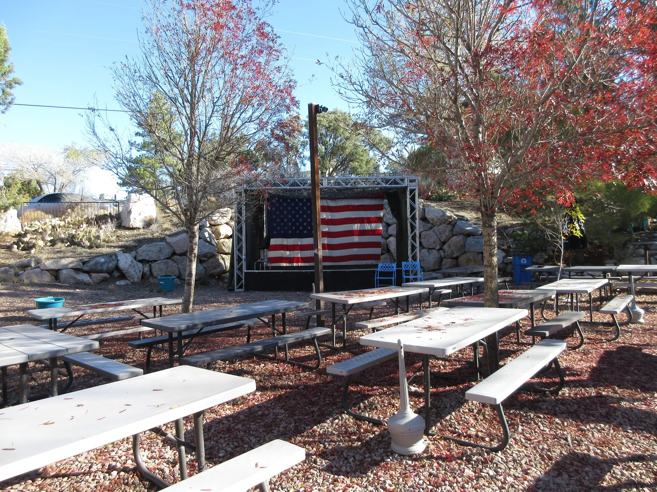 Outdoor seating area with picnic tables, trees with red leaves, a stage with an American flag backdrop, and a large rock wall.