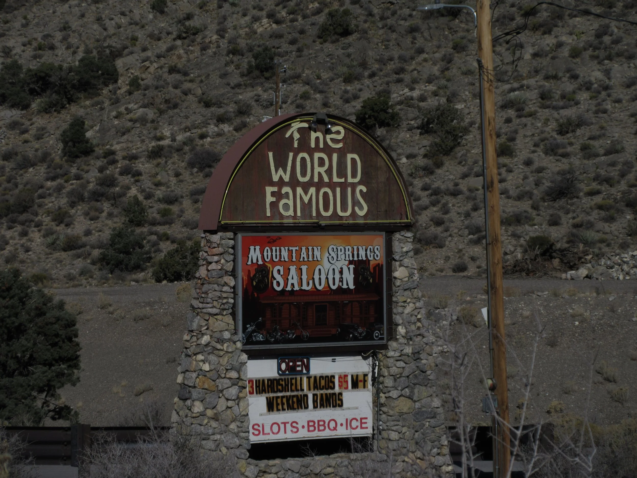 Sign for the World Famous Mountain Springs Saloon indicating it is open, with features including hard shell tacos, weekend bands, slots, BBQ, and ice, set against a desert landscape with hills and sparse vegetation.