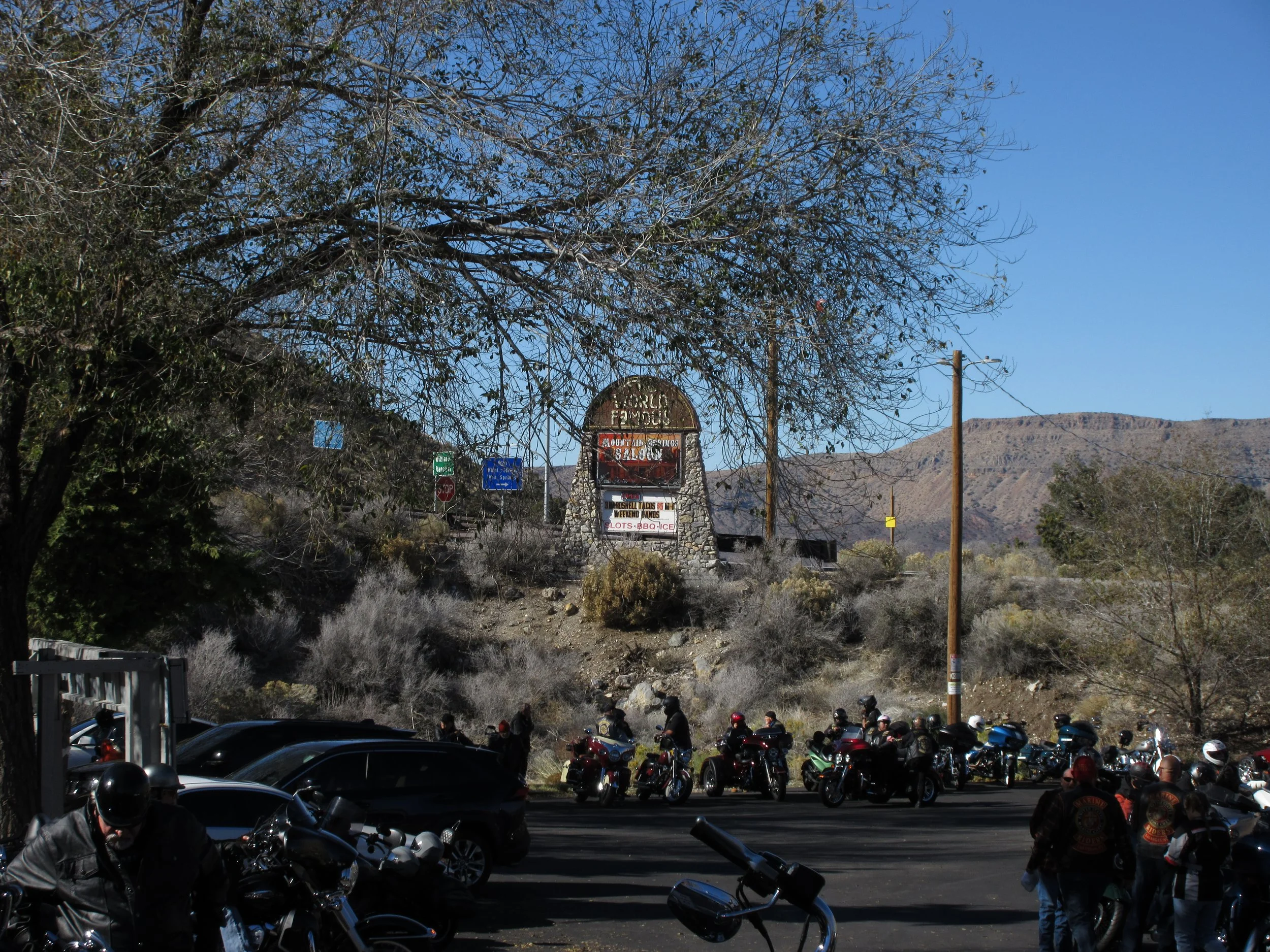 Motorcyclists gather near parked cars under a tree with a mountain landscape in the background. There is a sign reading 'Rough & Ready' and various traffic signs along the roadside.