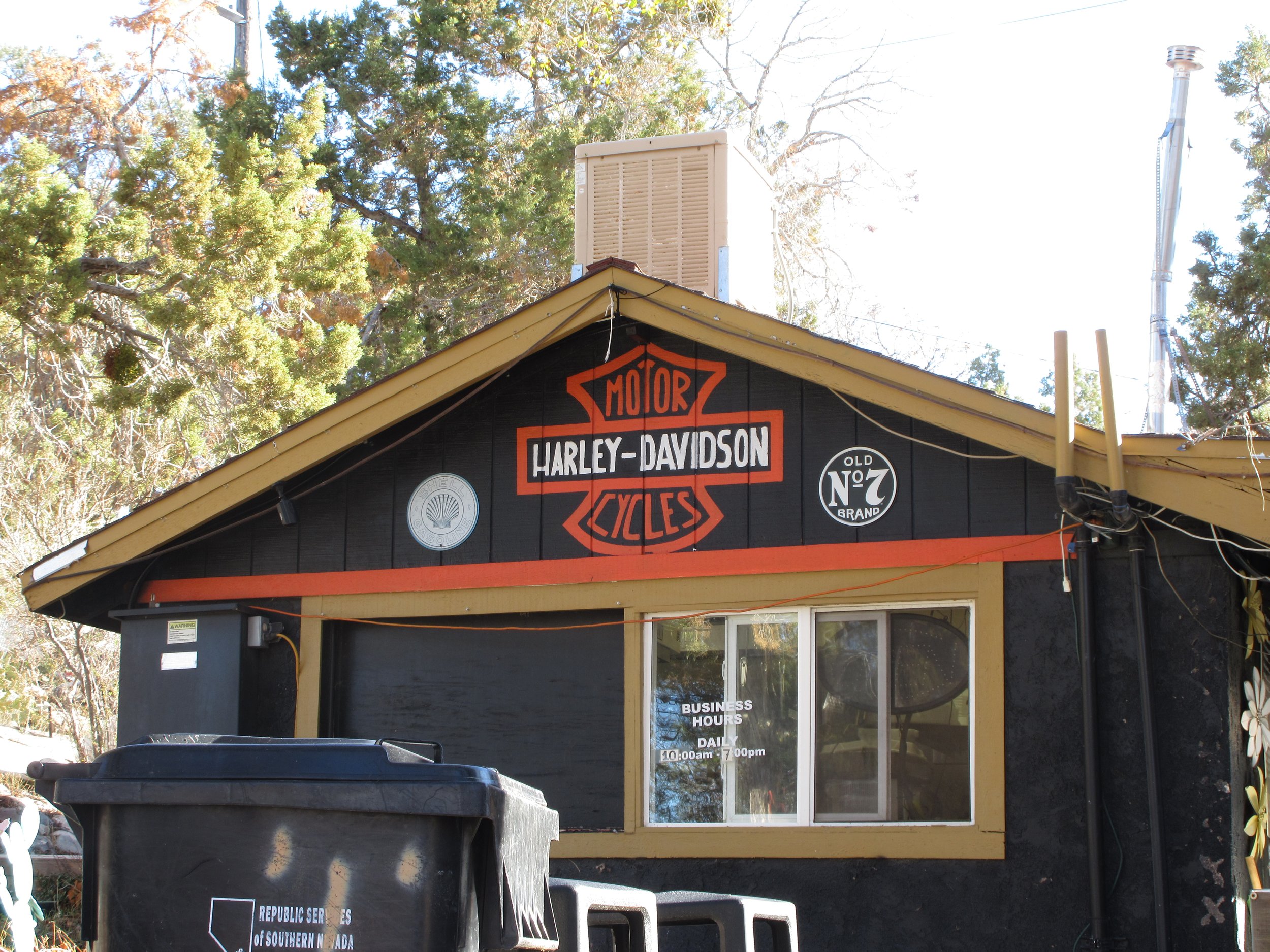 Harley-Davidson motorcycle shop with black exterior and yellow trim, displaying a sign with Harley-Davidson logo and old No. 7 brand logos, business hours are 10 am to 7 pm, surrounded by trees and a trash bin in the foreground.