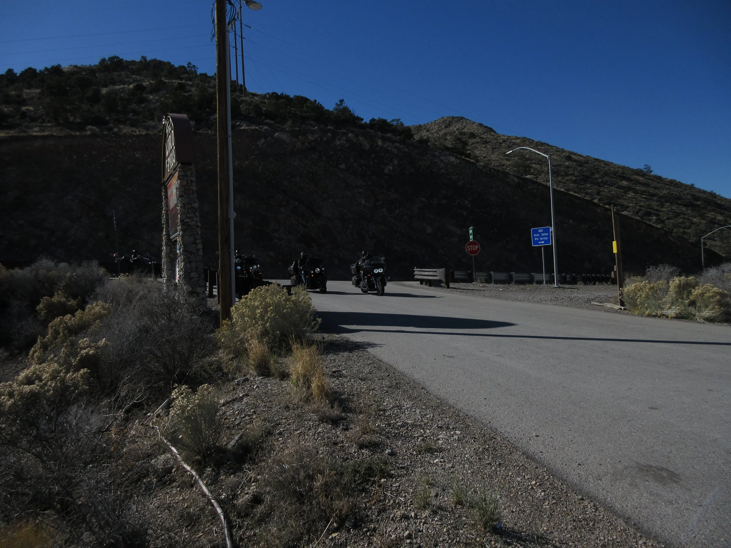 A small parking area on a mountain road with motorcycles sitting in a row. There are stop and road signs, and a large mountain in the background under a clear blue sky.