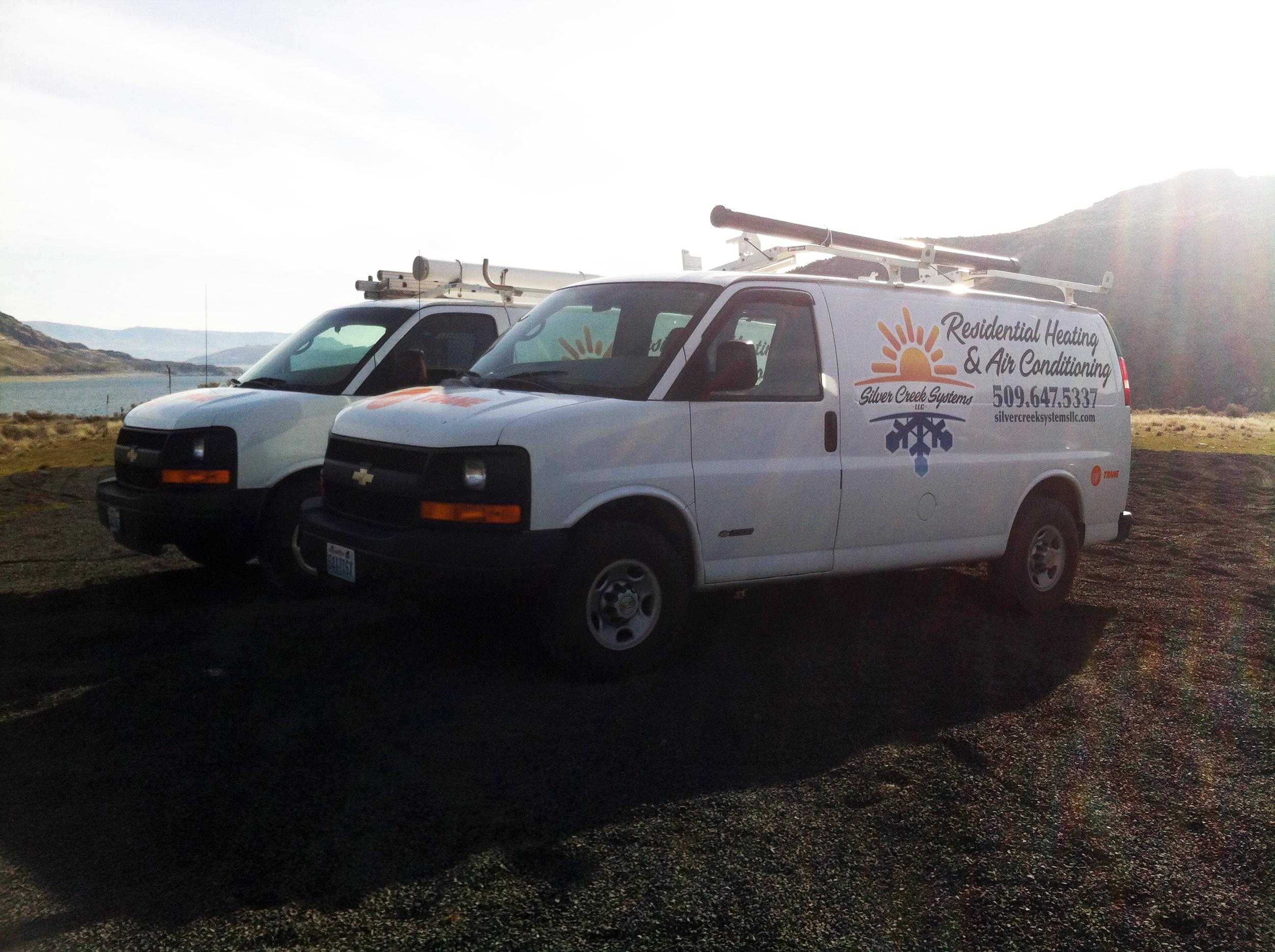 Two white service vans with company logo and contact information, parked outdoors with a mountain landscape and water in the background during sunset.