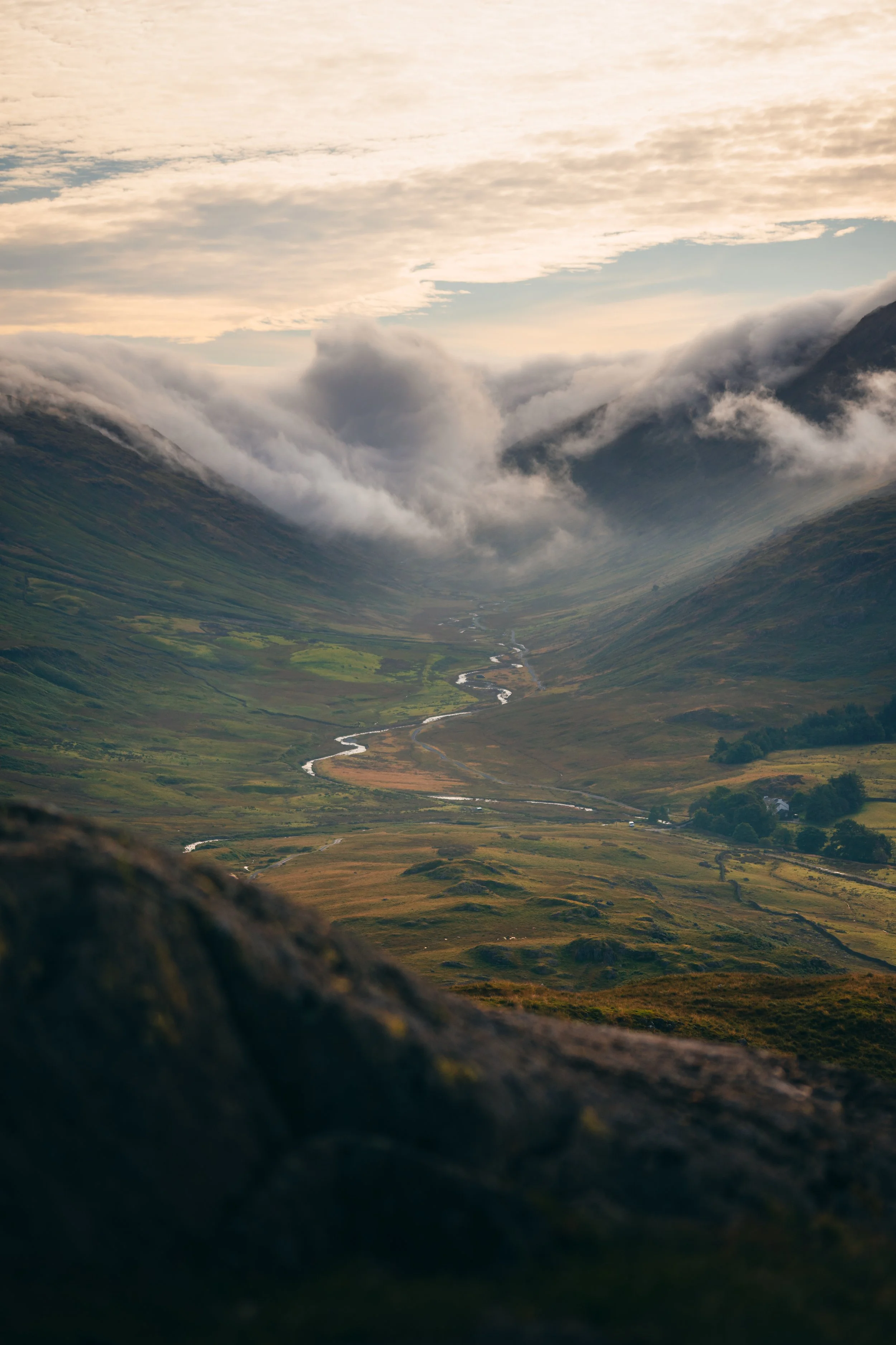 A scenic valley with a winding river, green fields, and mountains covered with clouds in the background, under a partly cloudy sky.