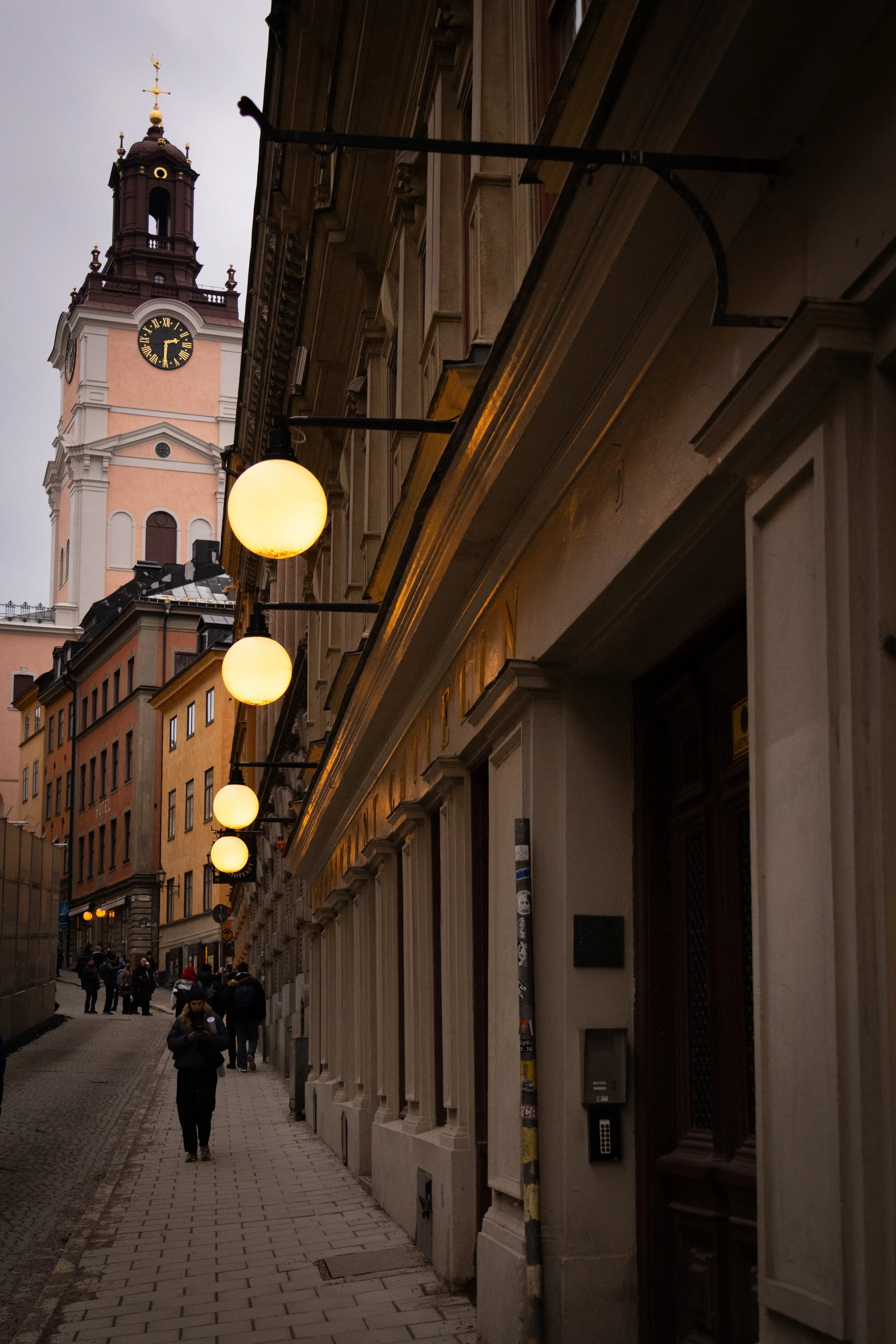 A city street at dusk with a tall clock tower in the background, lit spherical street lamps along the sidewalk, and people walking.