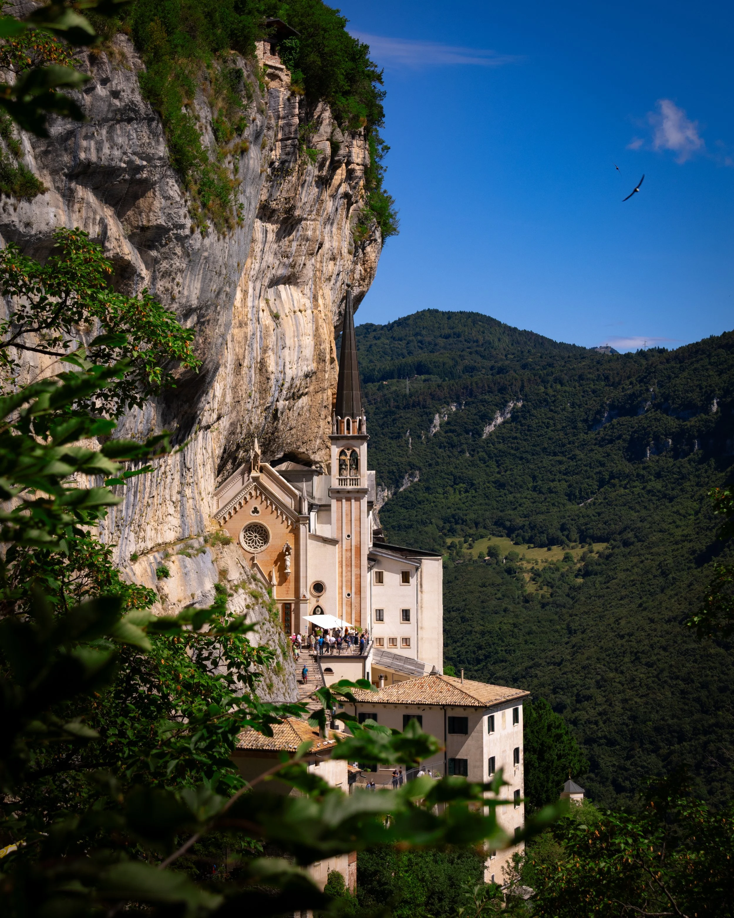 A church built into the side of a rocky hill with a steep, pointed steeple, surrounded by lush green hills under a bright blue sky with a few clouds and birds flying.