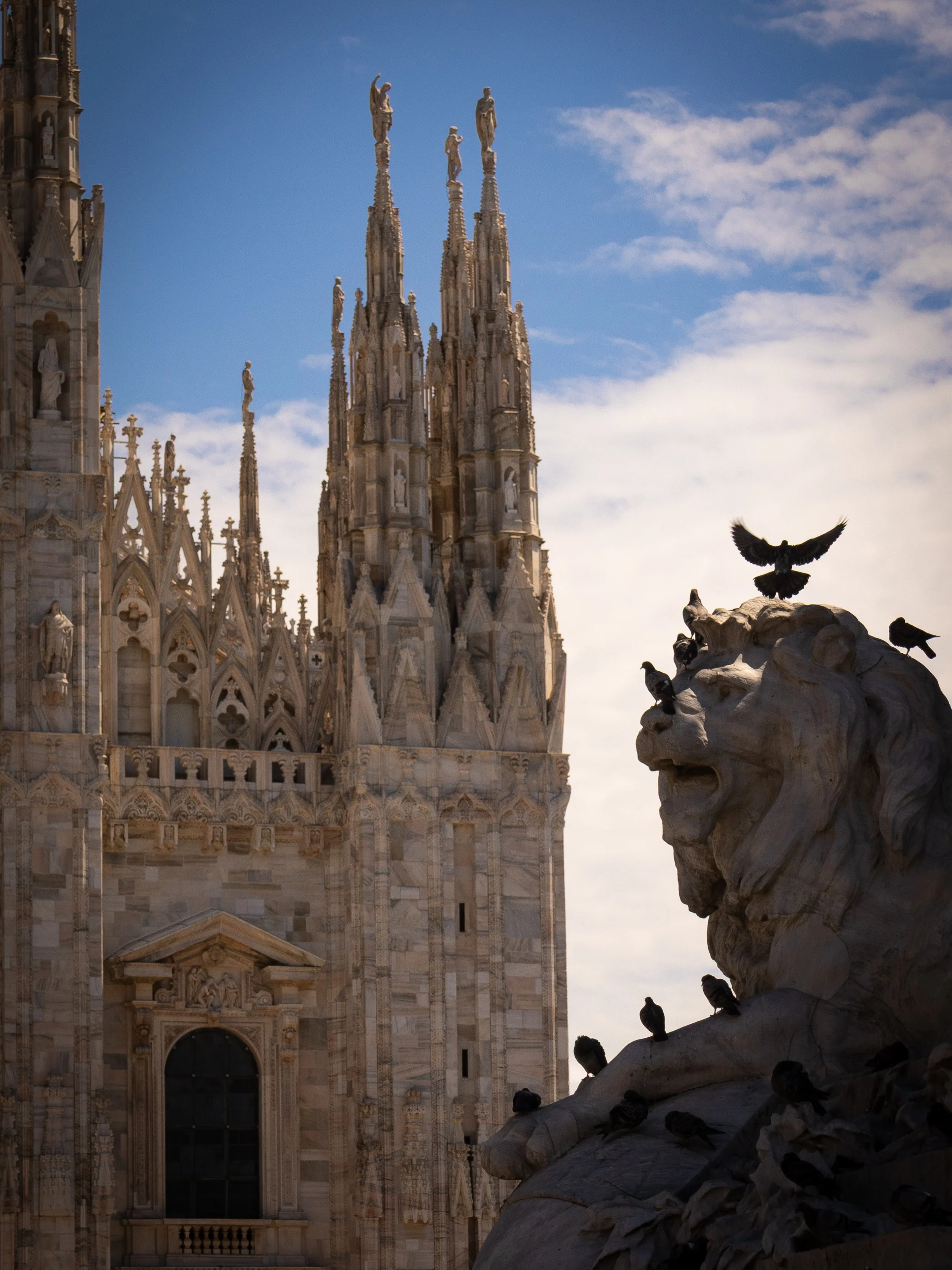 View of the Milan Cathedral with its intricate Gothic architecture and spires, and a marble lion statue with pigeons on and around it in the foreground, against a partly cloudy sky.