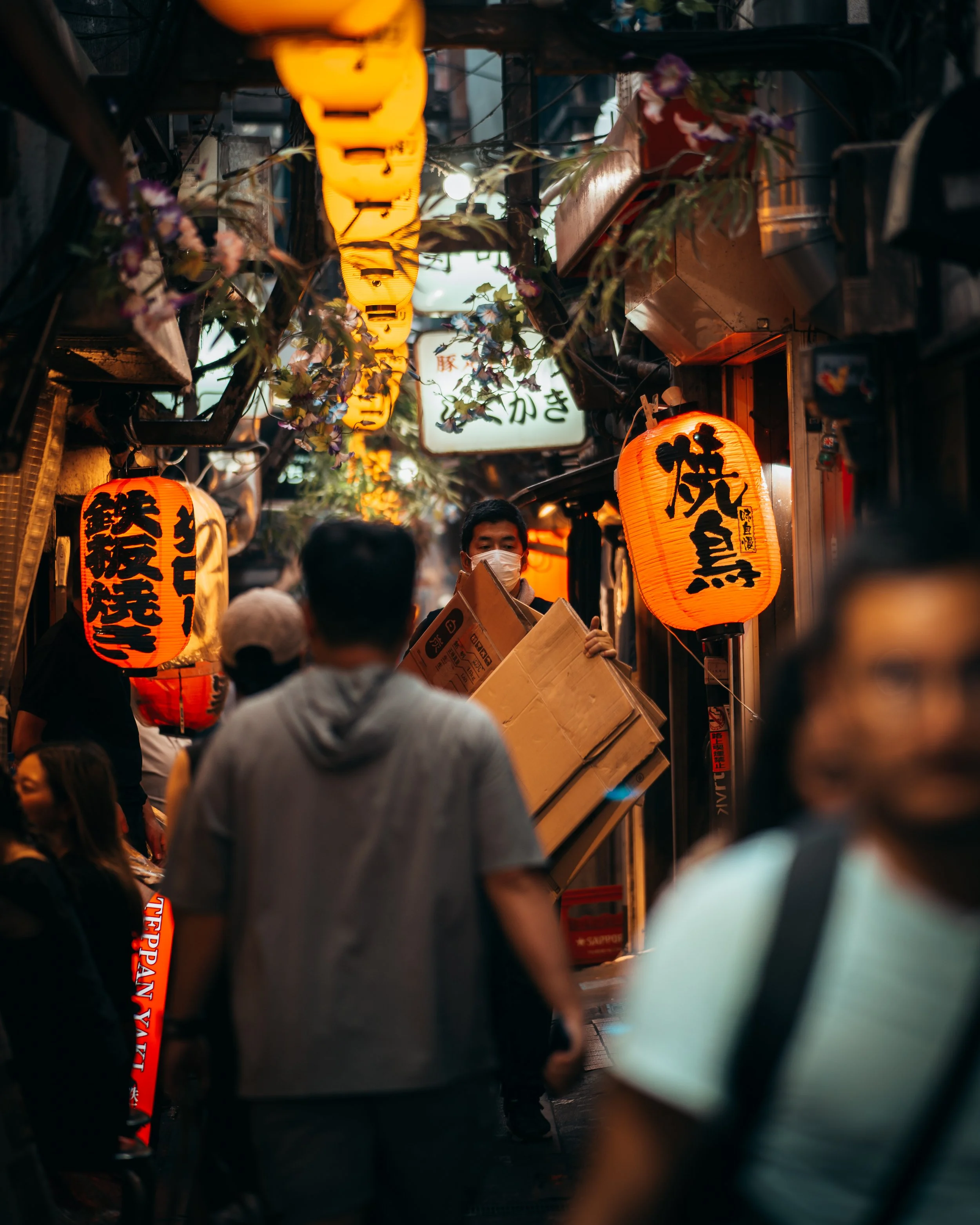 Night market street scene with orange and yellow lanterns, crowded with people, some wearing masks, narrow alley with shops displaying signs in Japanese, some people holding boxes and engaging in shopping or browsing.