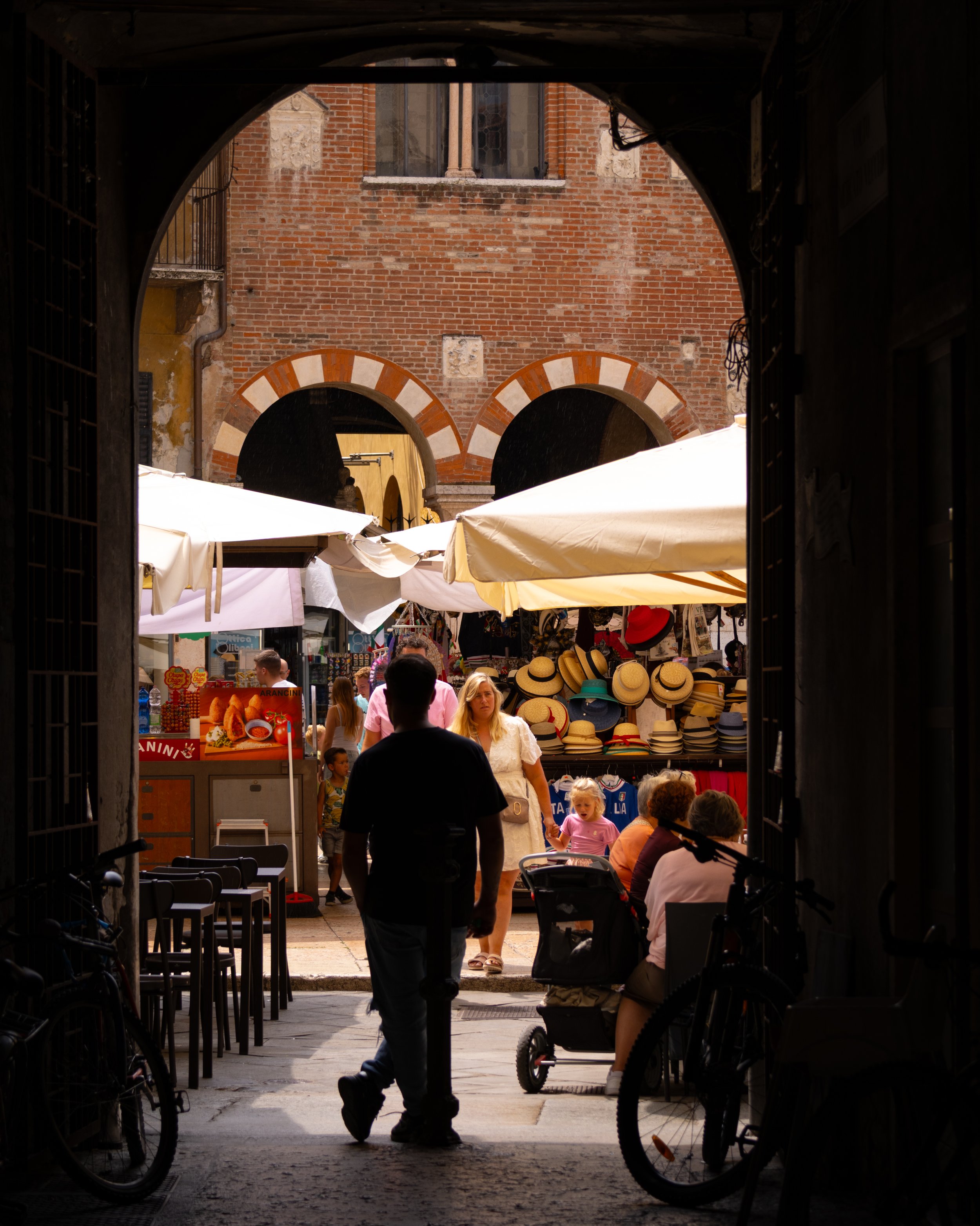 View of a marketplace through an arched entrance, with people shopping under tents and stall displaying hats, with red brick buildings in the background.